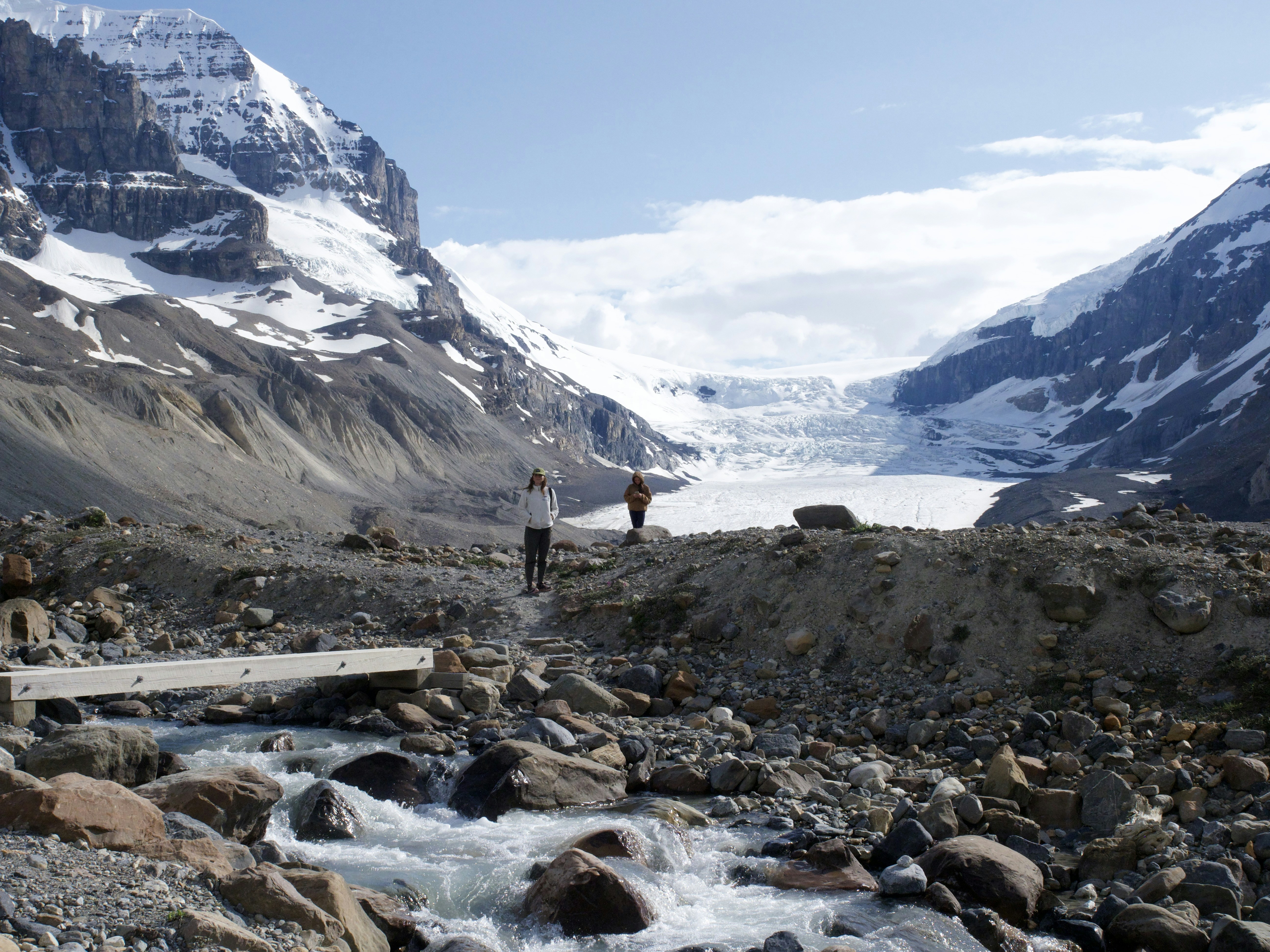 Due persone si trovano vicino a un ghiacciaio con montagne e ruscello.