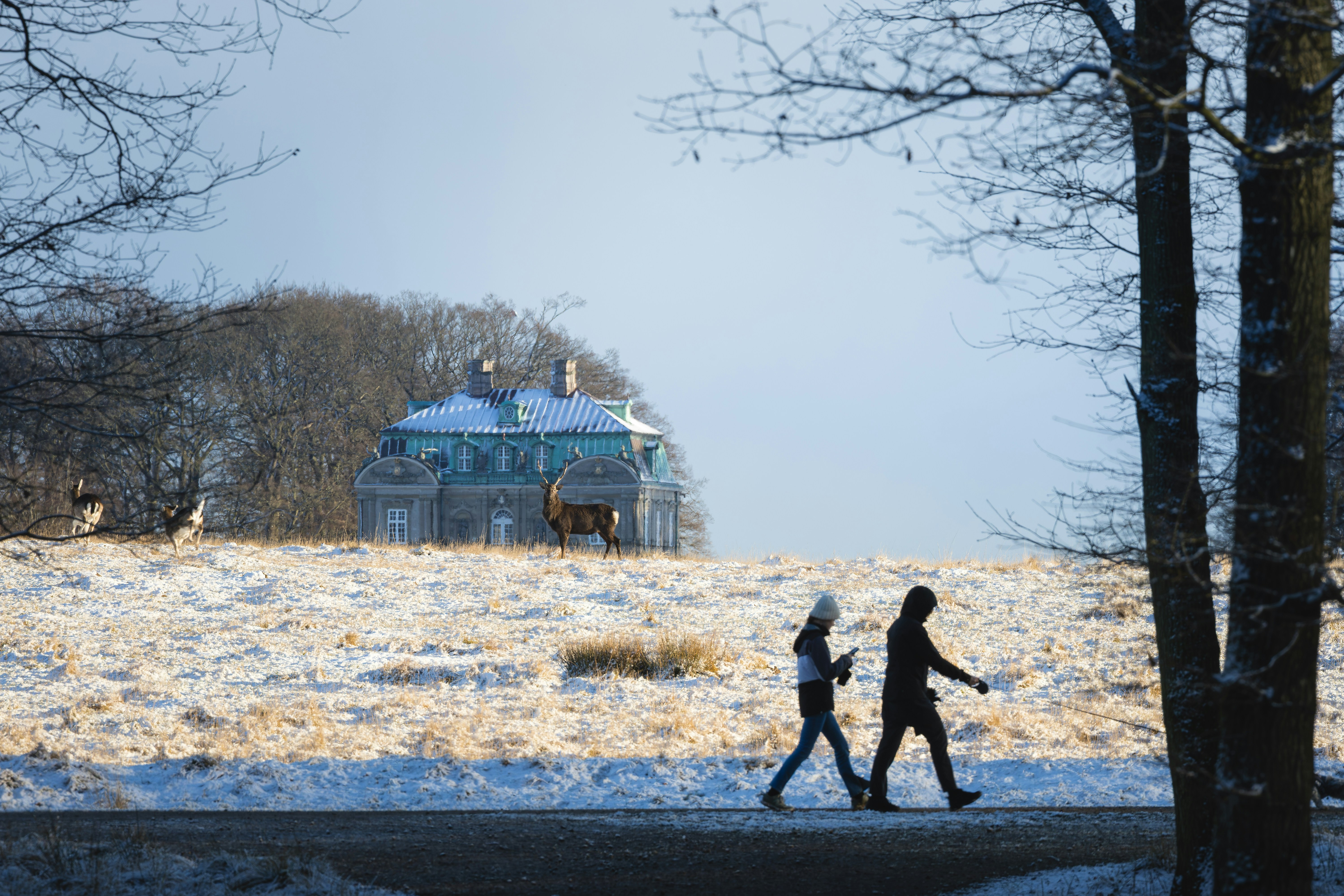 Two people walk past a snowy hill with a building.