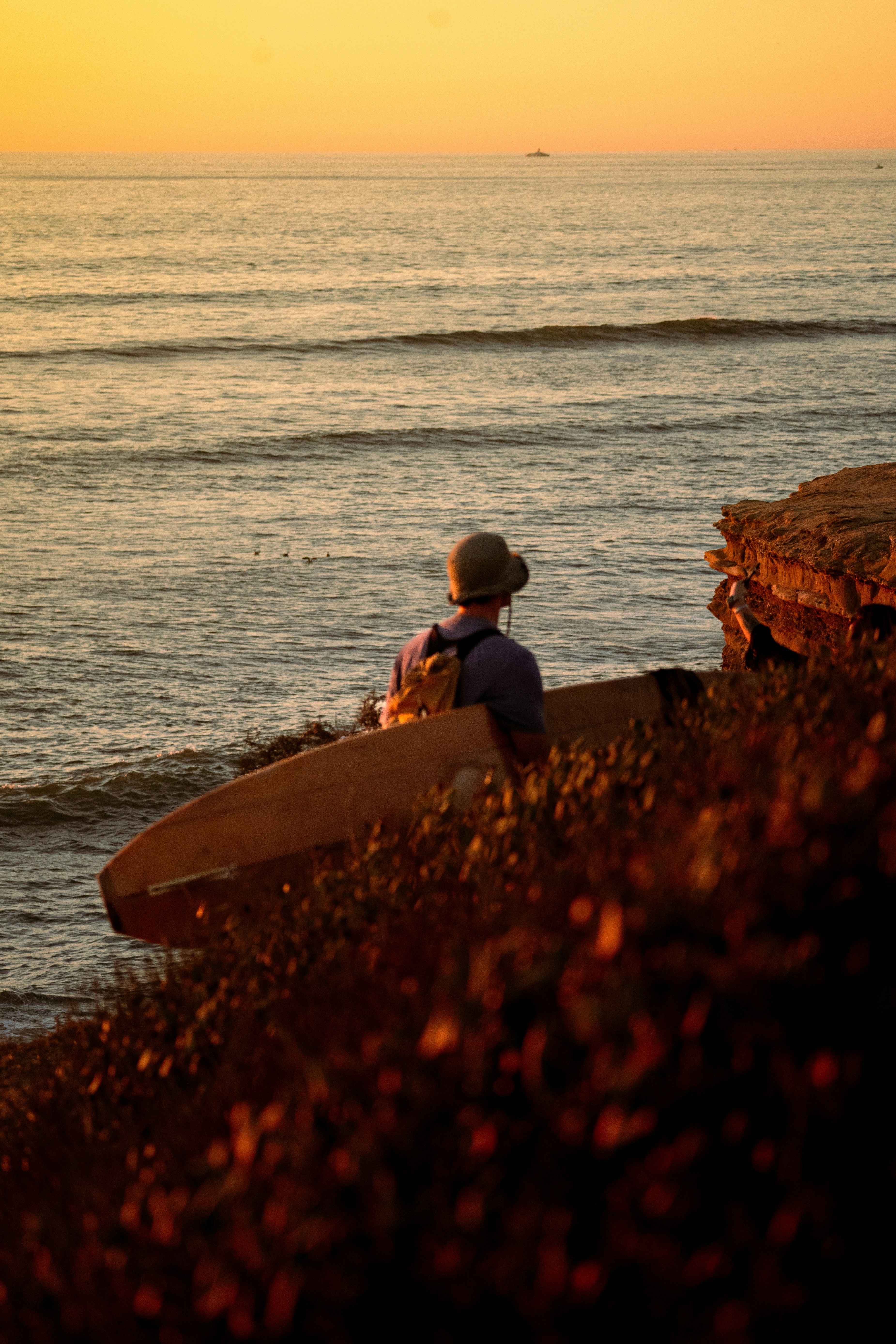 Surfer with surfboard on cliff overlooking ocean at sunset.