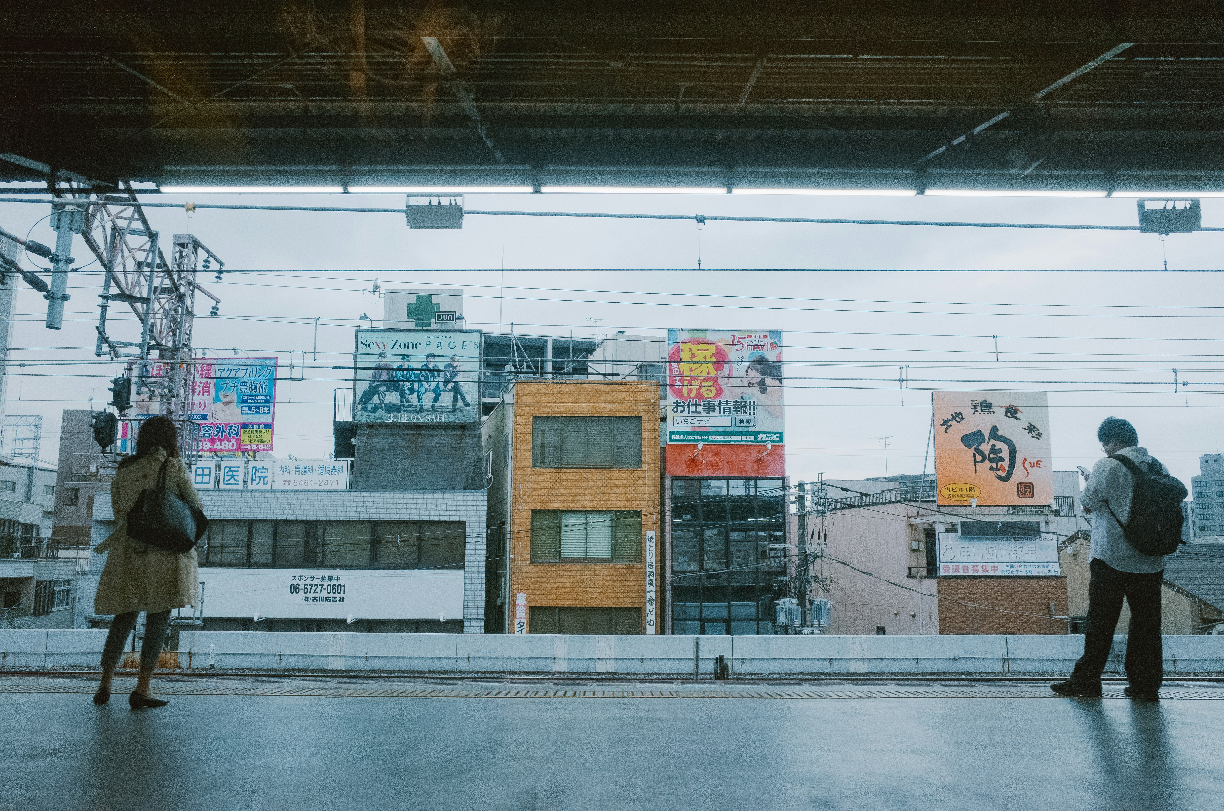 Due persone stanno su una banchina ferroviaria con edifici dietro.