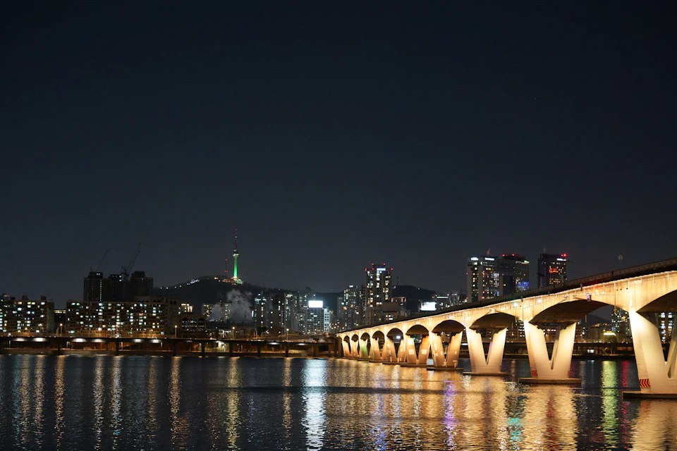 The image depicts a nighttime cityscape with a large, illuminated bridge spanning across a river. The bridge is adorned with lights that cast a warm glow on its white pillars and arches. In the distance, several tall buildings rise against the dark sky, their silhouettes adding depth to the scene. A green tower stands out among these structures, possibly serving as a landmark or a point of interest for residents and visitors alike. The calm waters below reflect the lights from the bridge and the surrounding cityscape, creating a beautiful interplay of light and shadow.