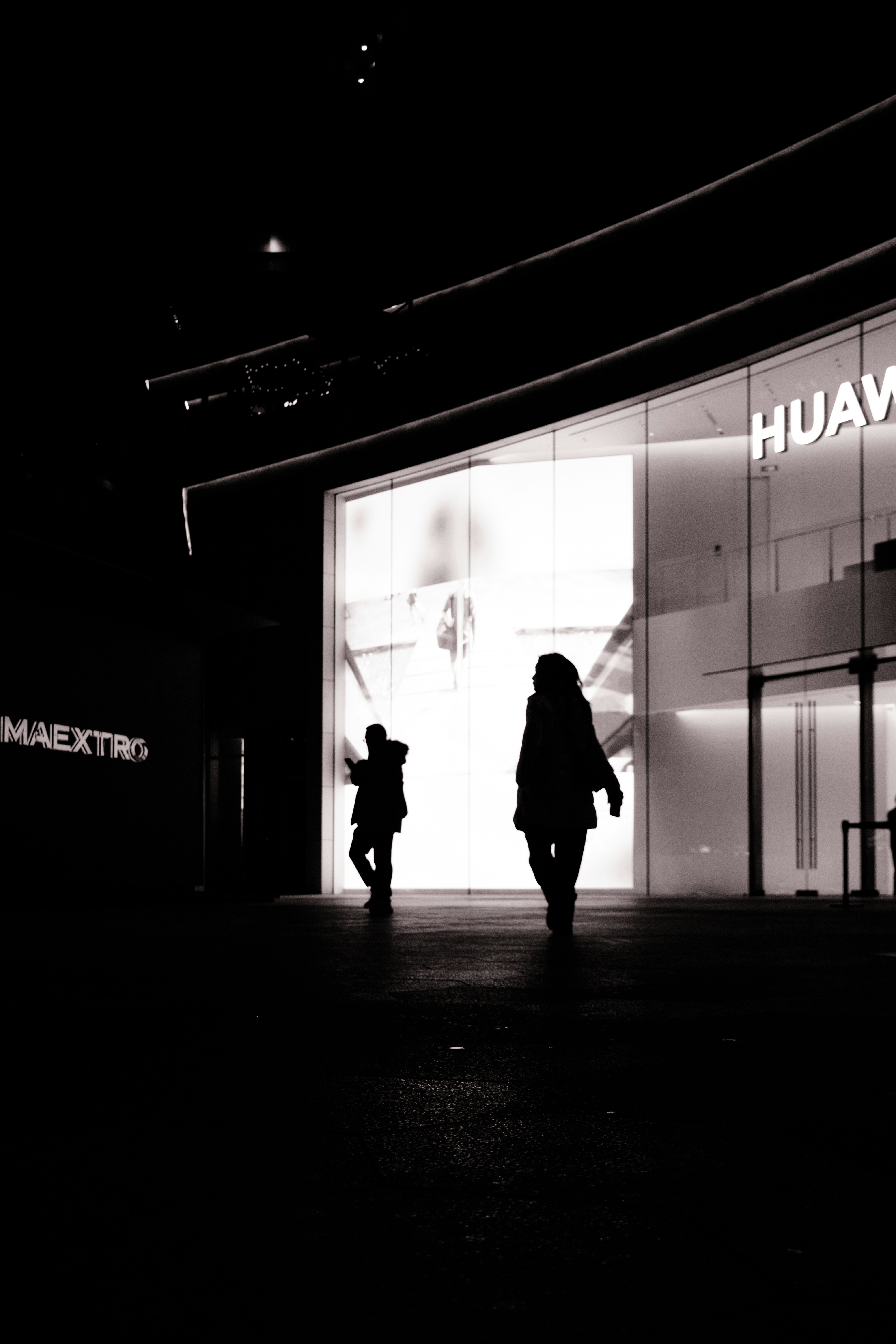 Two silhouettes walk towards a brightly lit building at night.