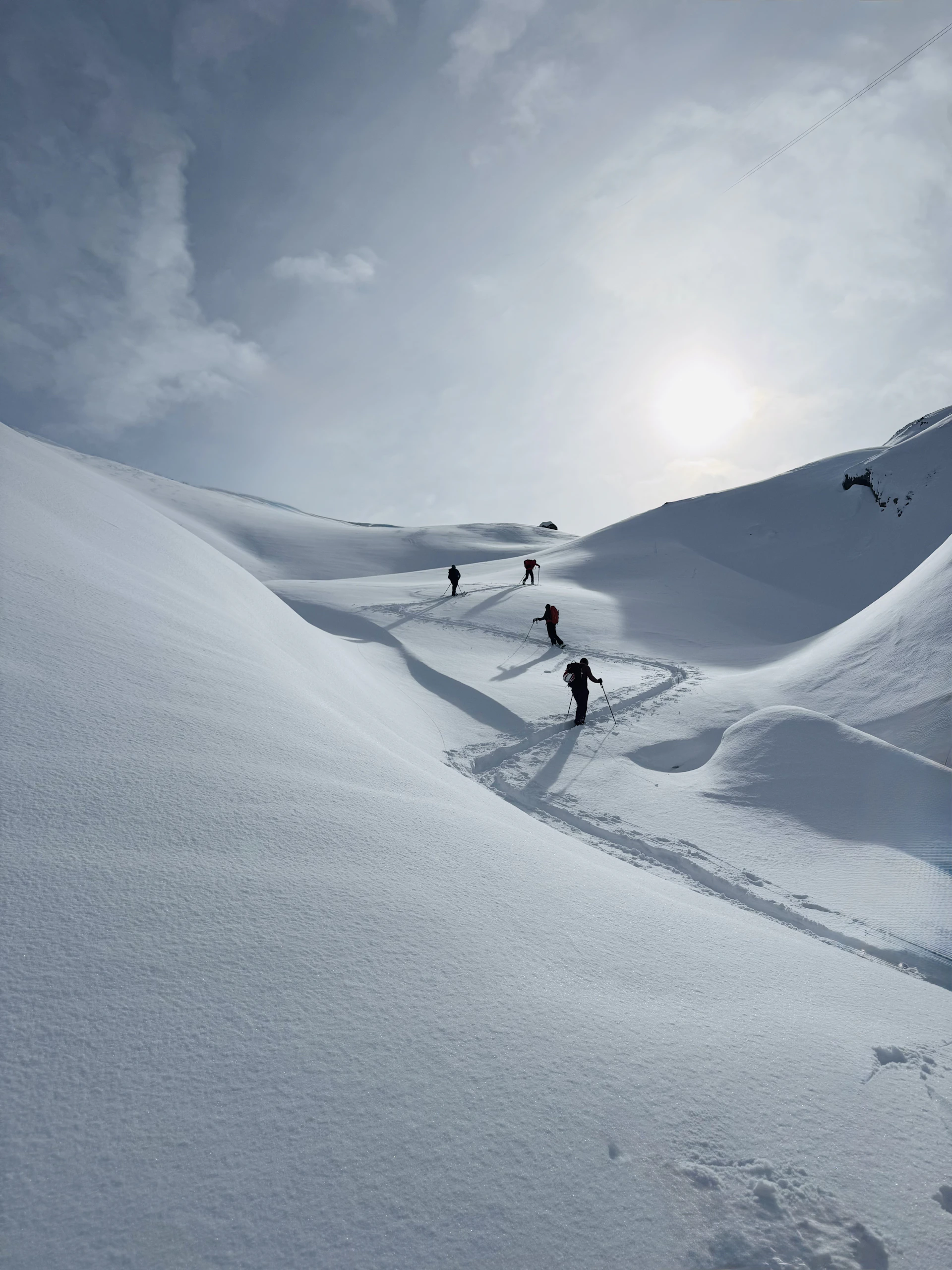 Skiers ascend a snowy mountain path under a bright sun.
