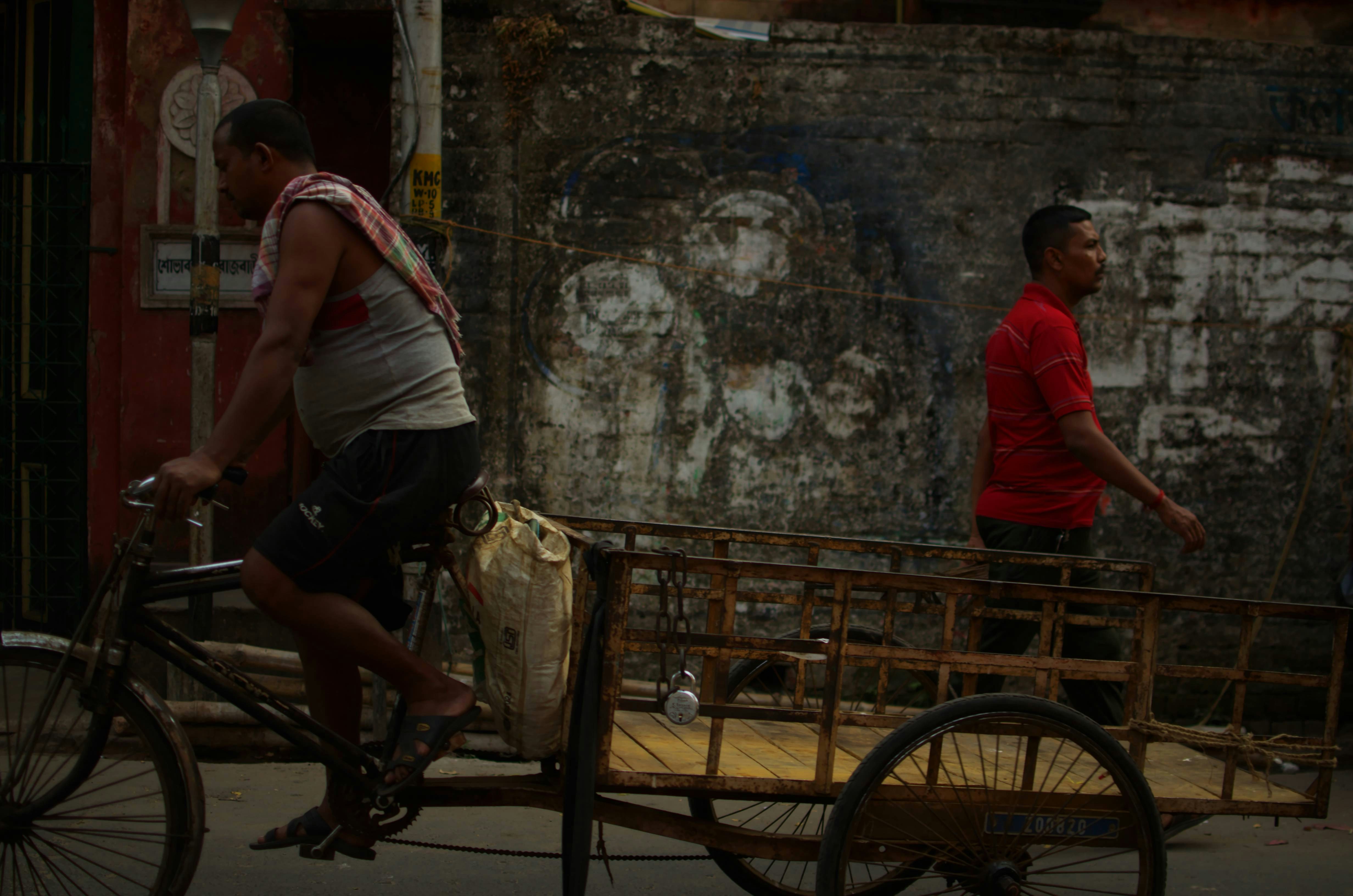 Man rides bicycle with cart past weathered wall.