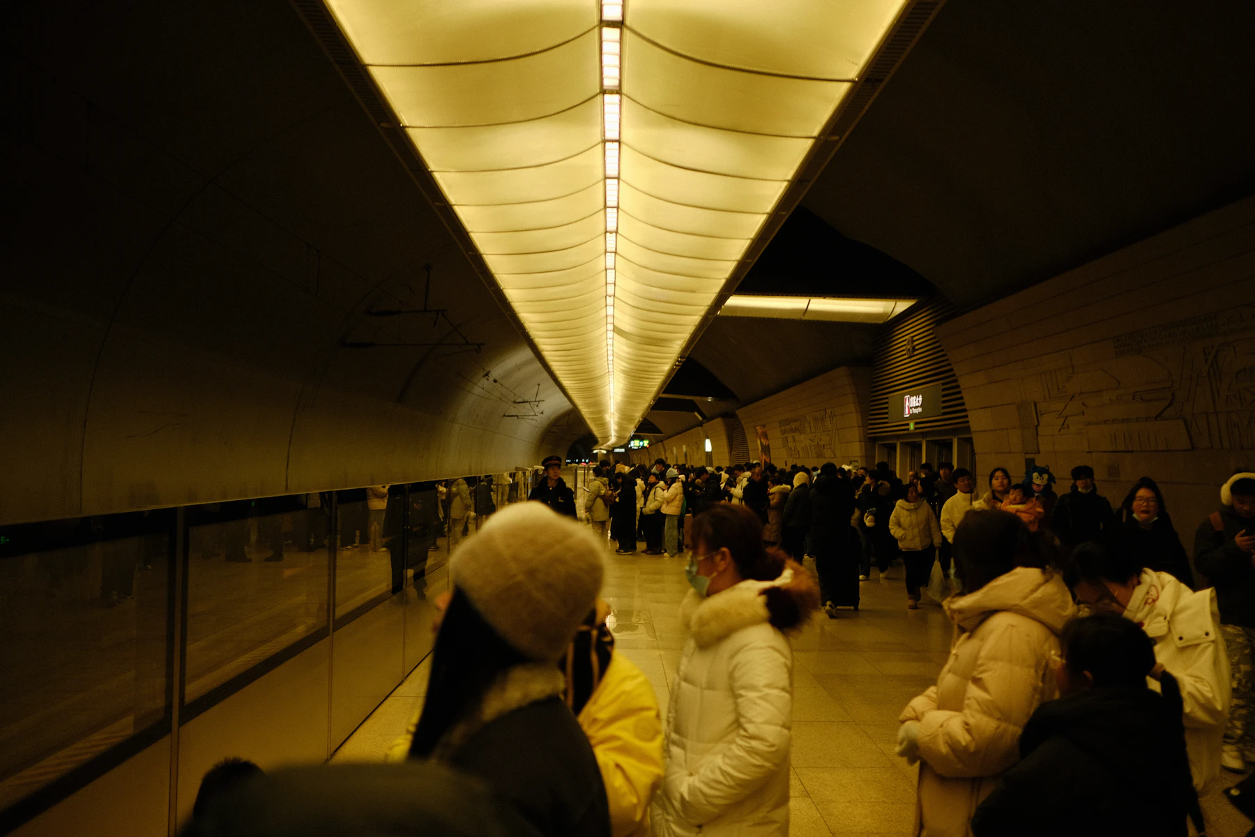 Travellers gather along a brightly lit underground railway platform.