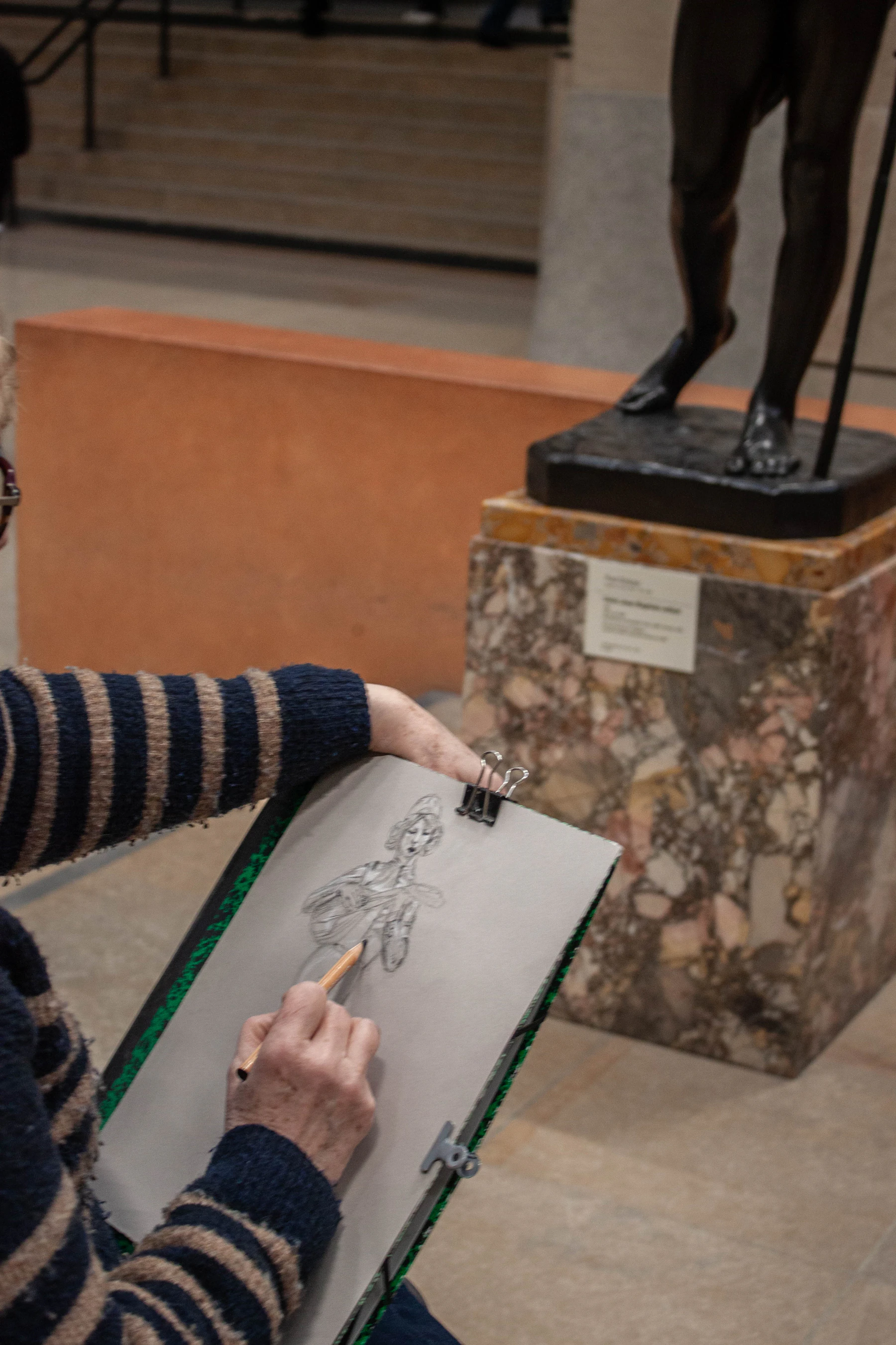 A visitor's hands drawing a bronze statue in pencil on a sketchpad, with the statue on its stone plinth just beyond