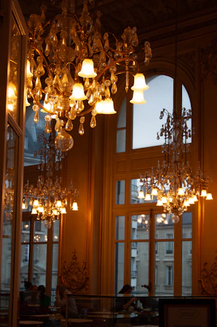 Ornate chandeliers illuminate a grand room with large windows.