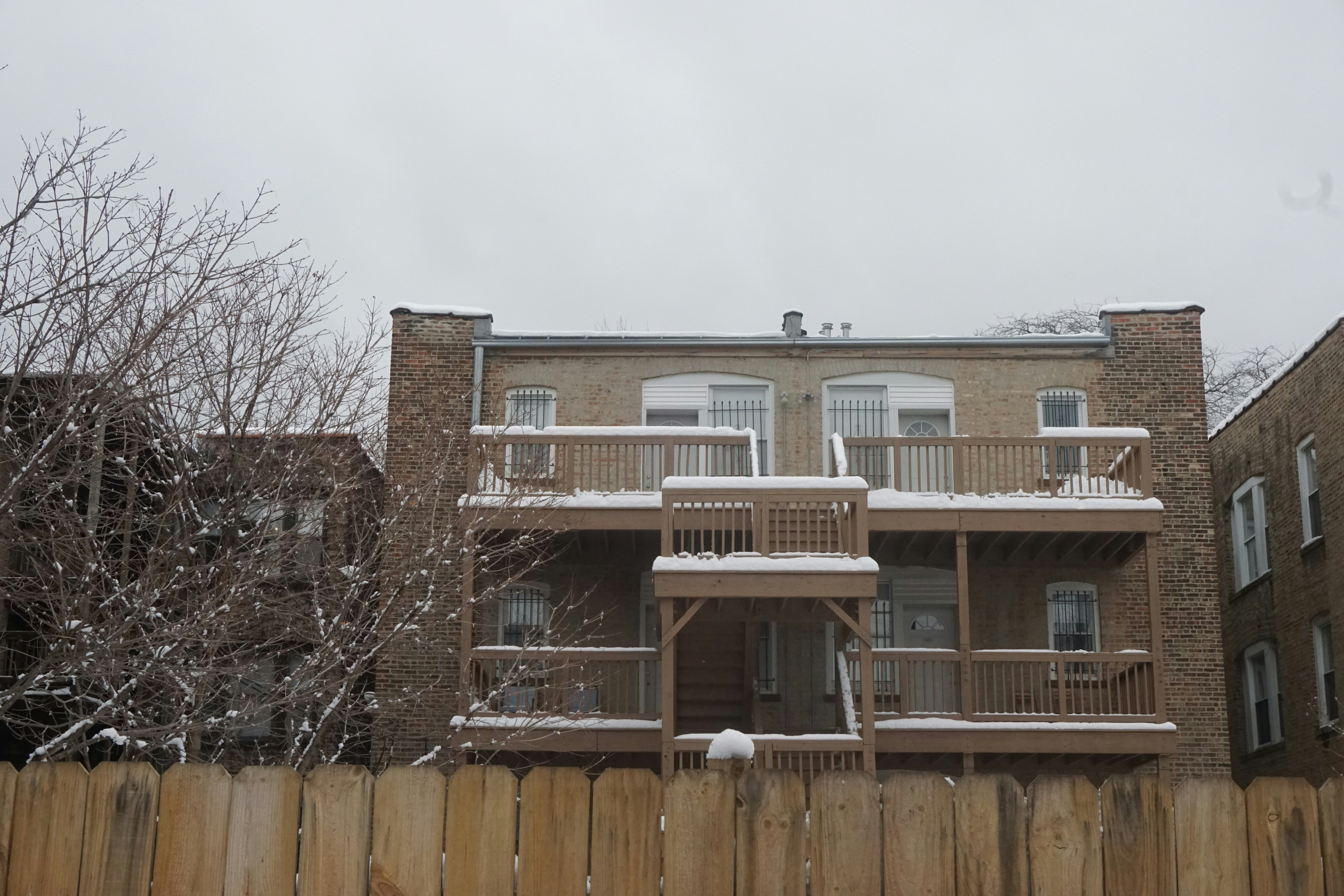 A brick building with tiered wooden decks covered in snow is seen over a wooden fence, with bare trees under a cloudy sky.