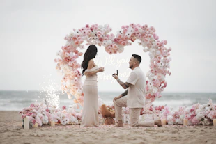 Man proposes to woman on beach with heart arch.