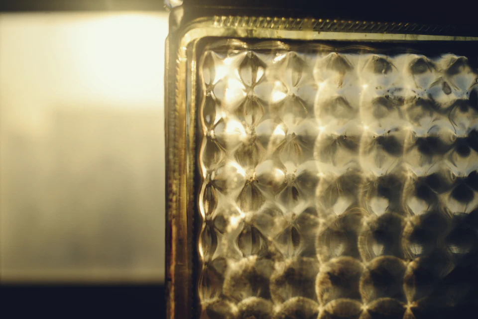 The image shows a close-up view of a glass surface, likely a window or mirror, with a metallic sheen to it. The glass appears to be made of metal and has a reflective quality that catches the light. The background is blurred, drawing focus to the glass surface in the foreground.