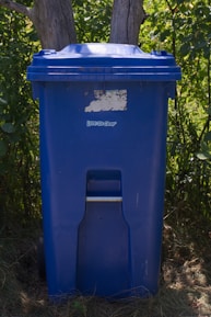 A blue trash can sits outdoors near trees.