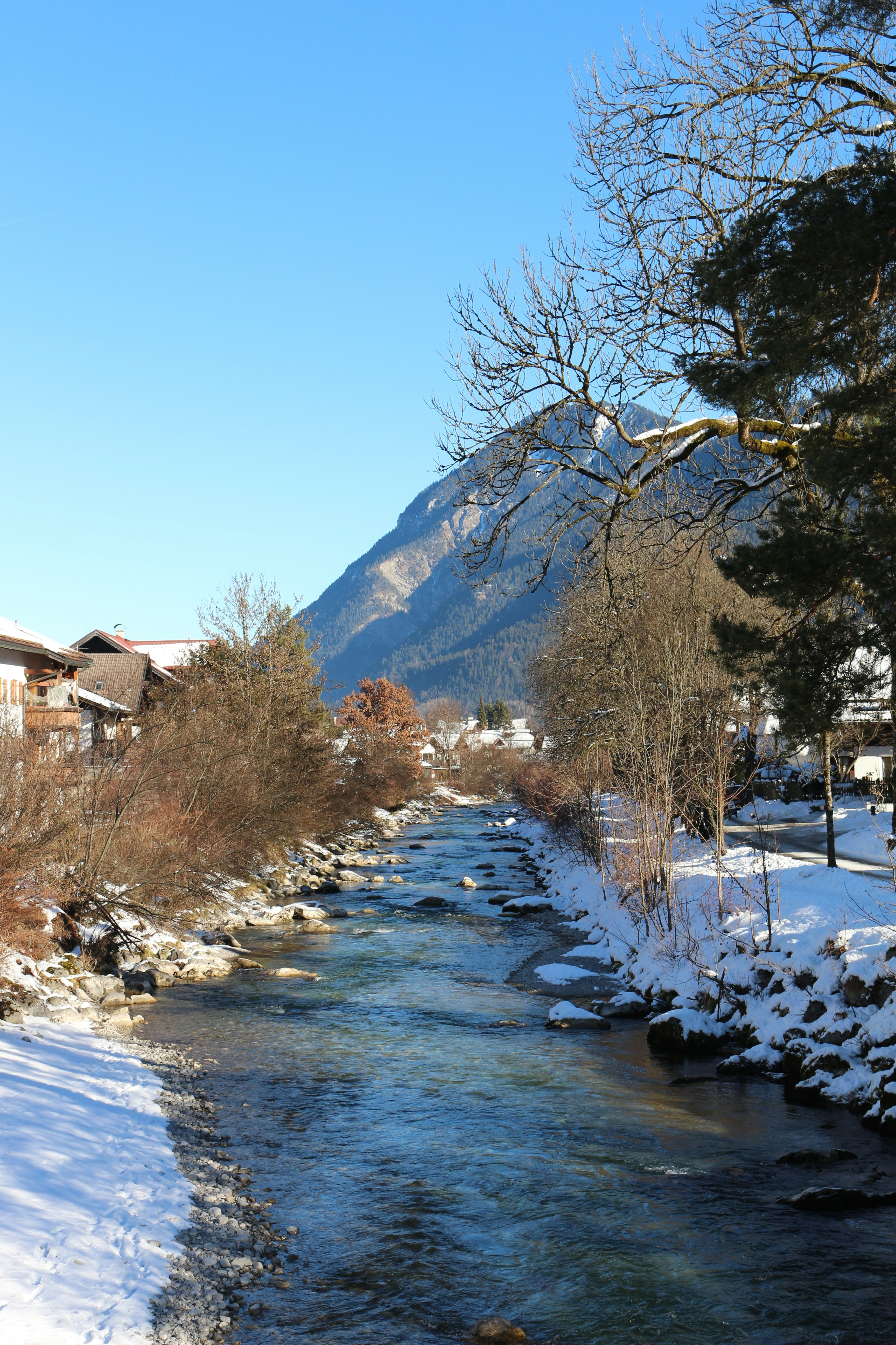 A clear river flows through a snowy village with mountains.