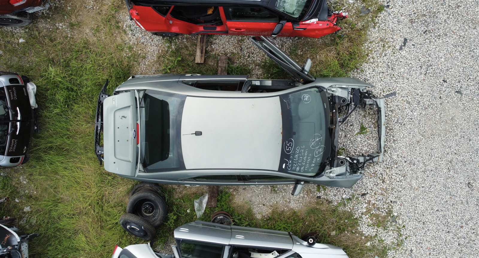Aerial of a stripped silver donor hatchback in the salvage yard, windshield marked for VIN and stock