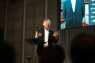 An elderly asian man speaks animatedly on a stage