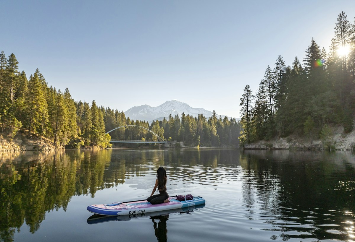 A person kneeling on a paddleboard on a still lake ringed by pine forest and a distant mountain