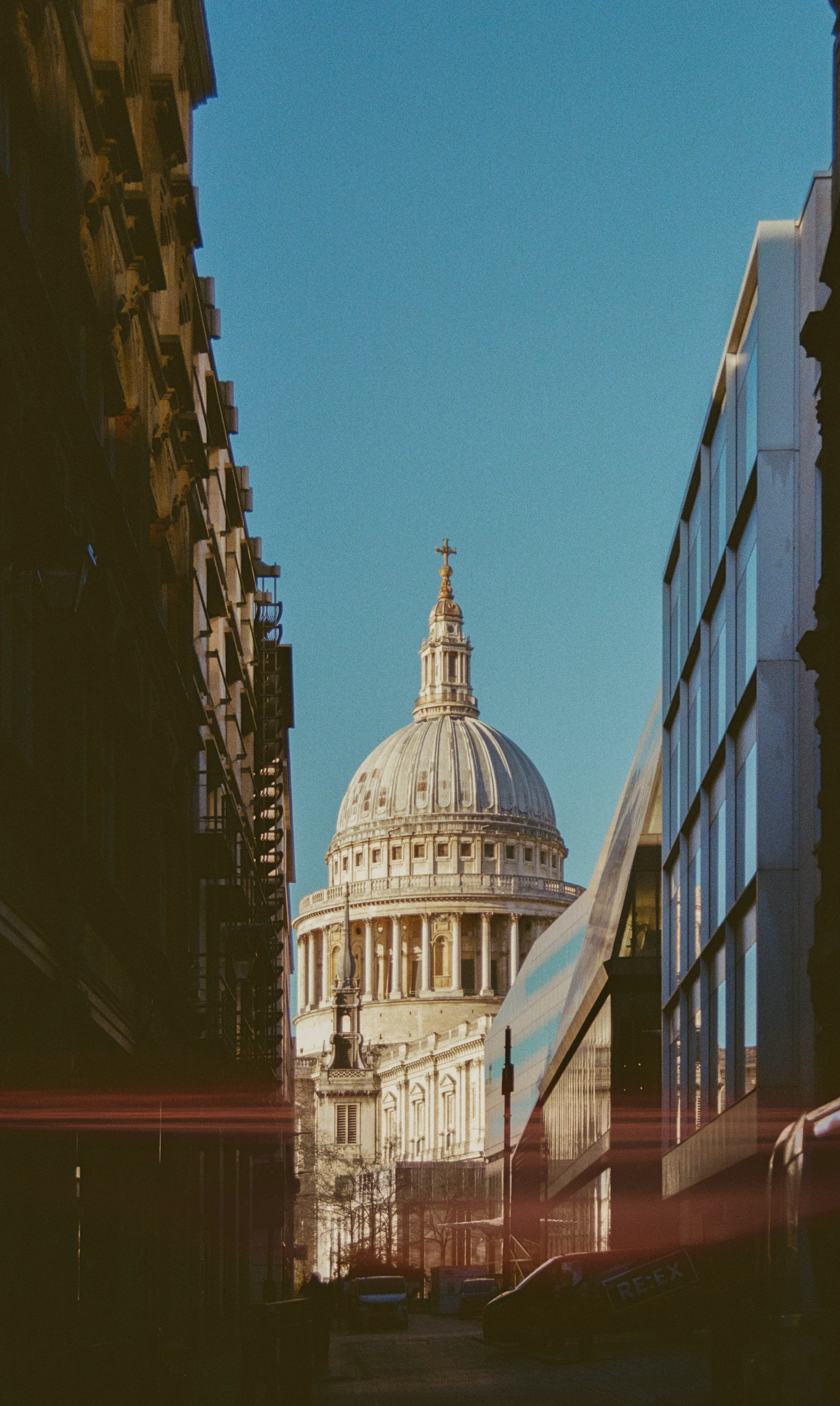 Photograph of The dome of St Paul’s Cathedral framed between brick and glass buildings on a narrow City street.