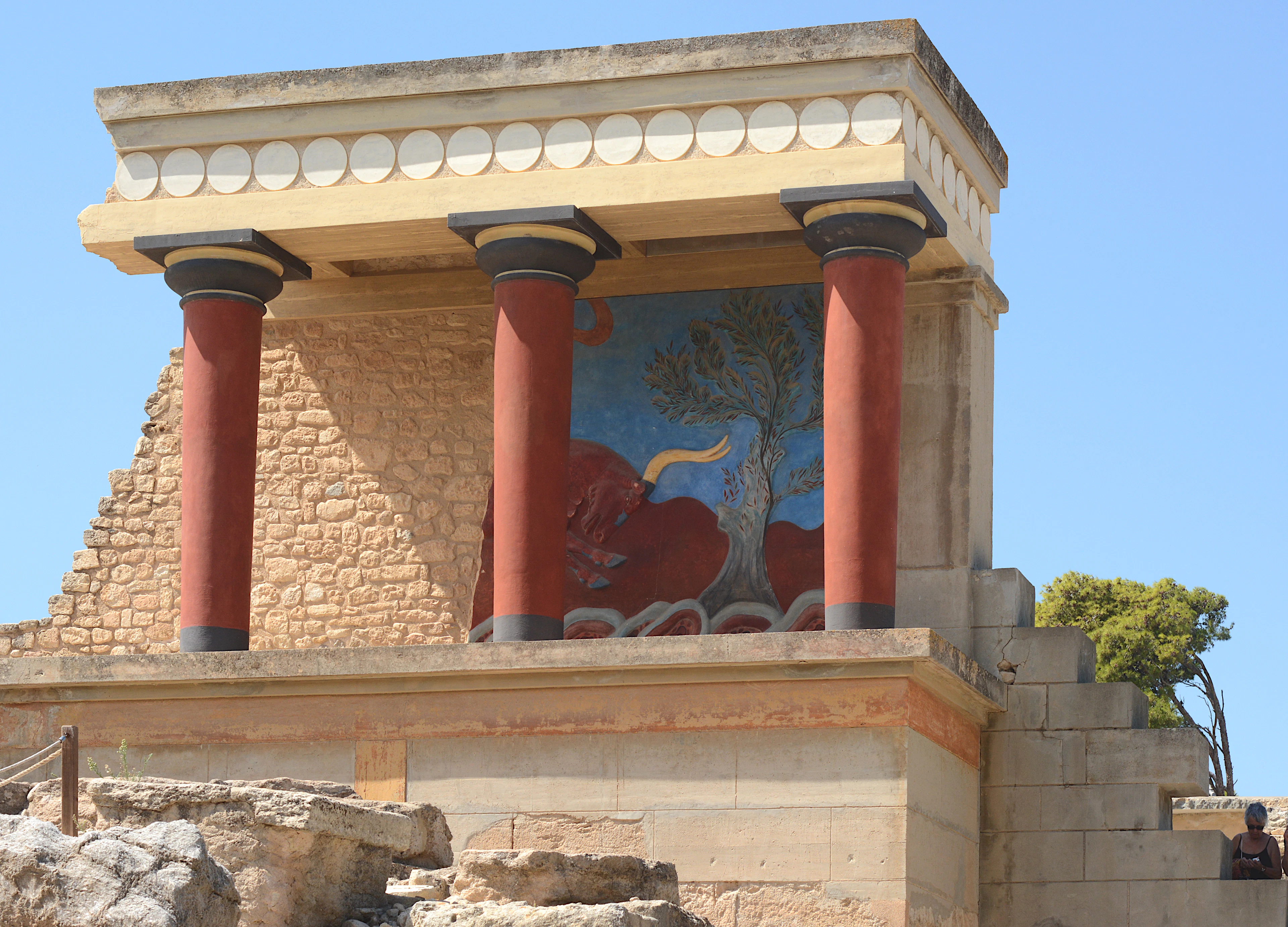The restored colonnade at the north entrance of the Minoan palace of Knossos on Crete: three tapered crimson columns beneath a reproduction of the charging-bull fresco, set against a deep blue sky