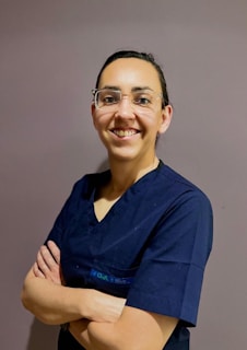 A woman in blue scrubs with glasses smiles.