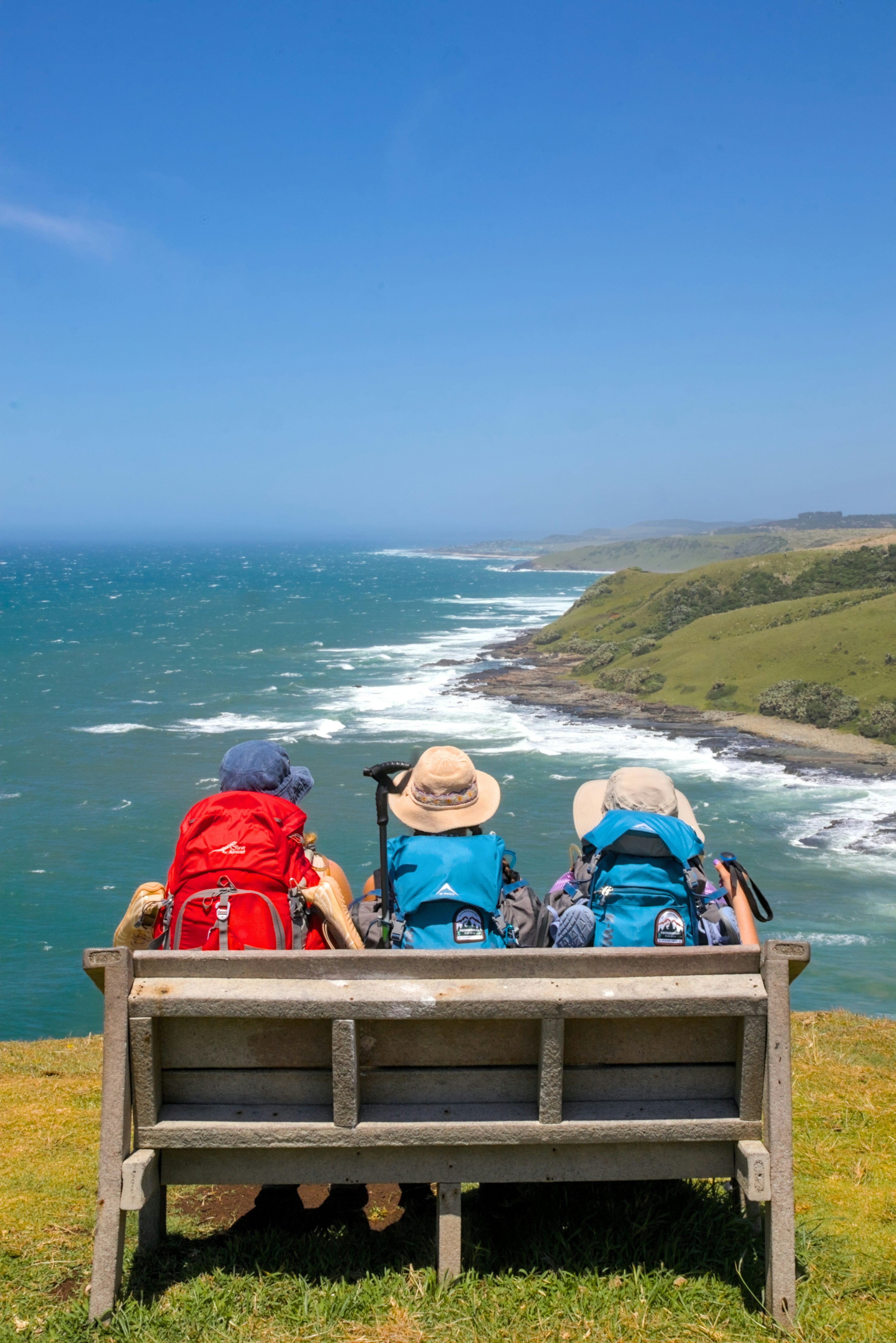 Three hikers with backpacks enjoy ocean view from bench