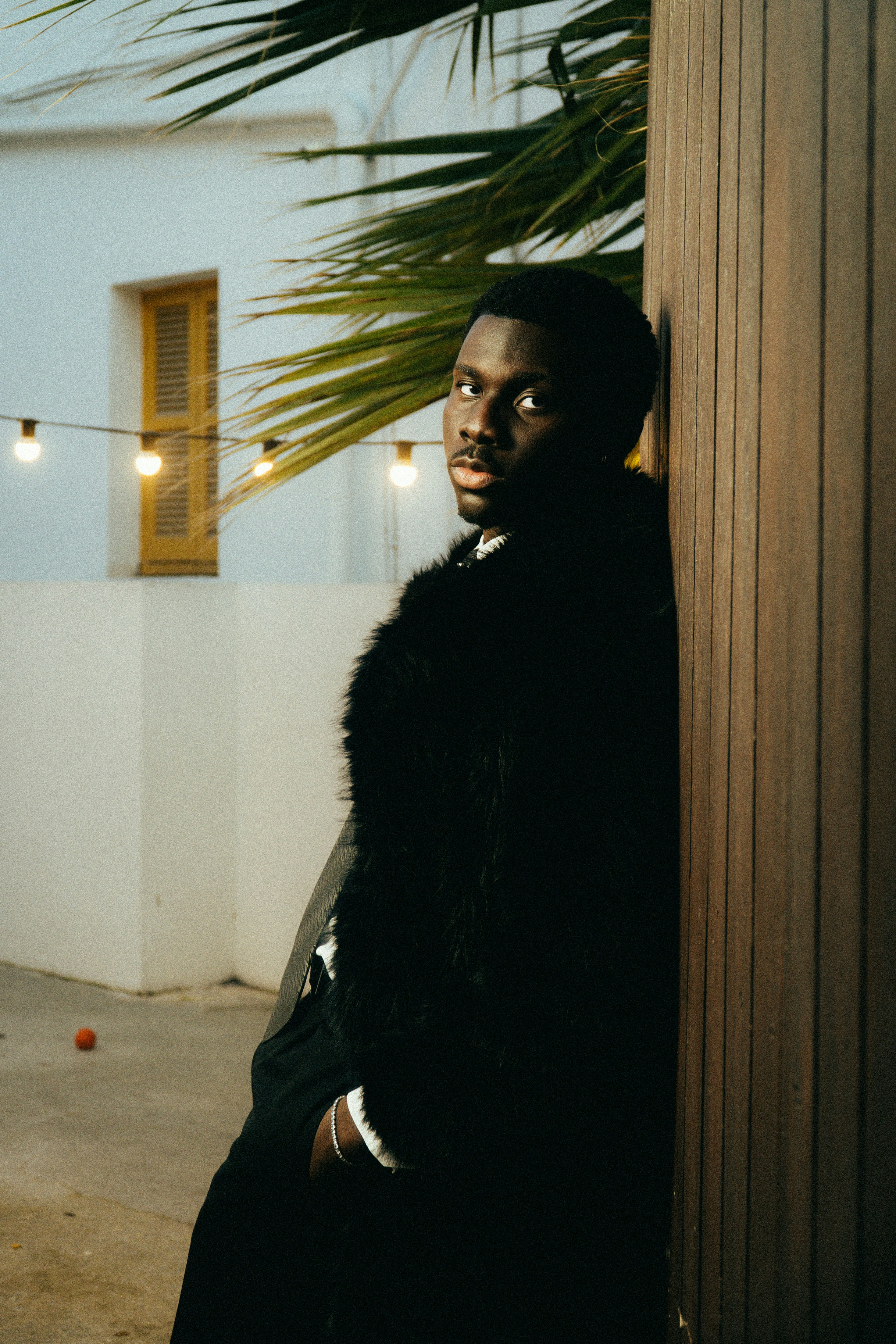 Man in fur coat leaning against wooden post outdoors.