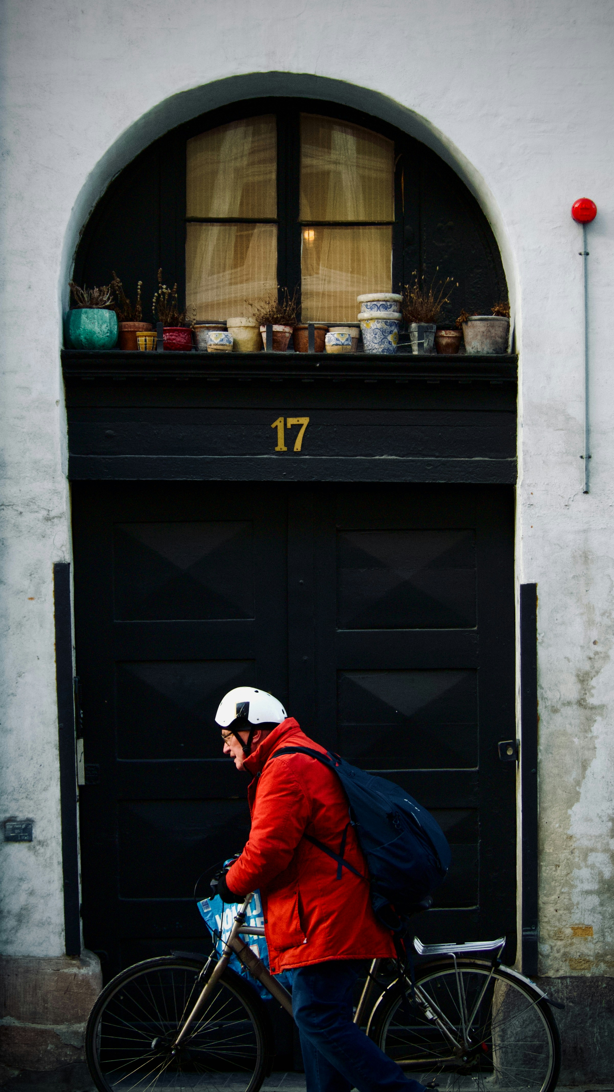 Man in red jacket rides bicycle past dark doorway.