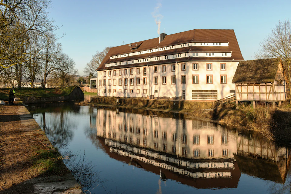 The image depicts a large, white building with a brown roof situated on the bank of a river. The building features numerous windows and appears to be a residential or commercial structure. A small bridge can be seen spanning across the river, connecting two parts of the land separated by water. The sky above is clear blue, suggesting a bright and sunny day.