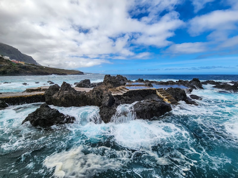 The dramatic natural volcanic pools of El Caletón in Garachico, Tenerife, with Atlantic waves crashing against the dark lava rocks under a cloudy sky.
