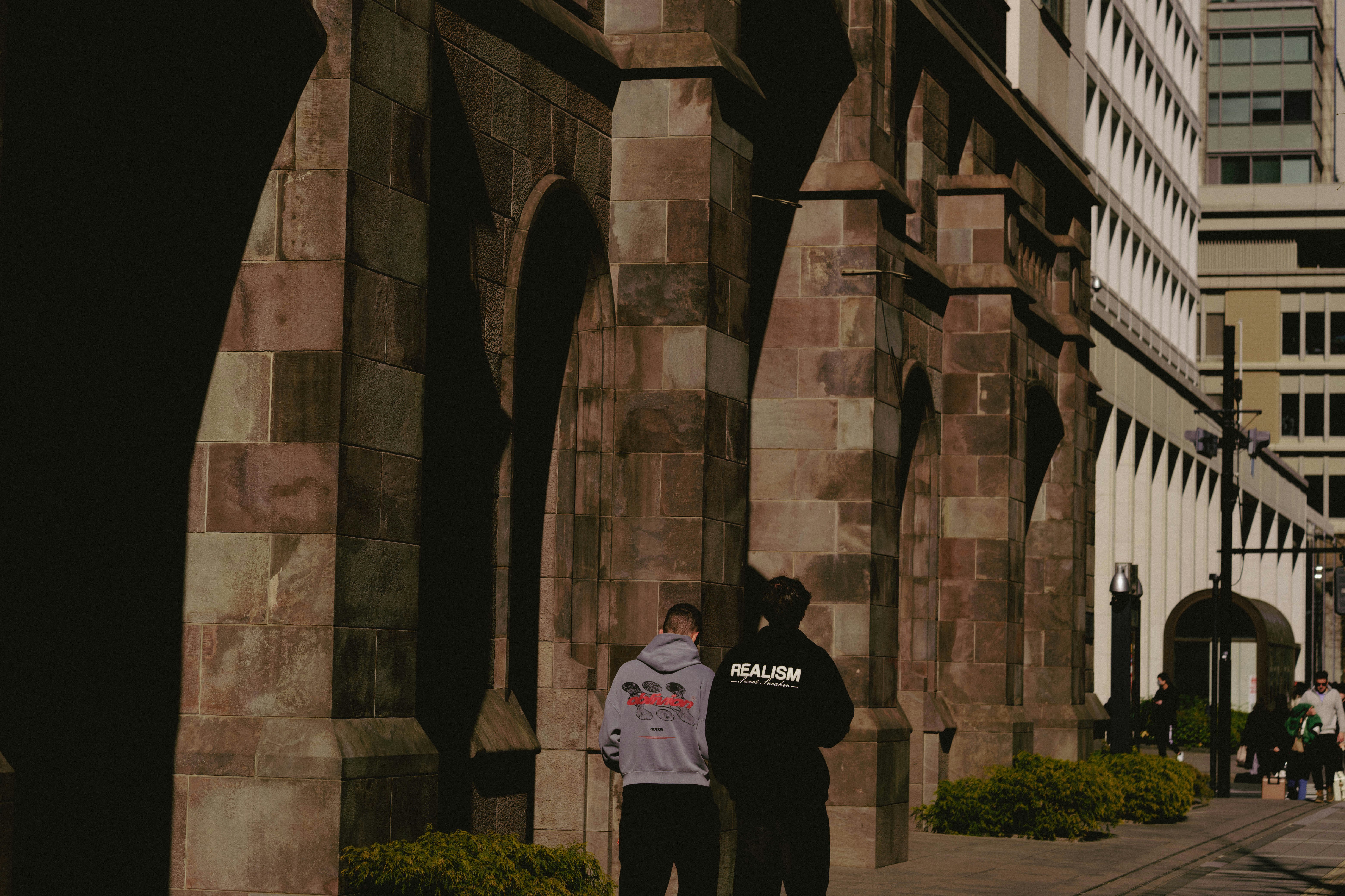 Two people walk past an old stone building with arches.