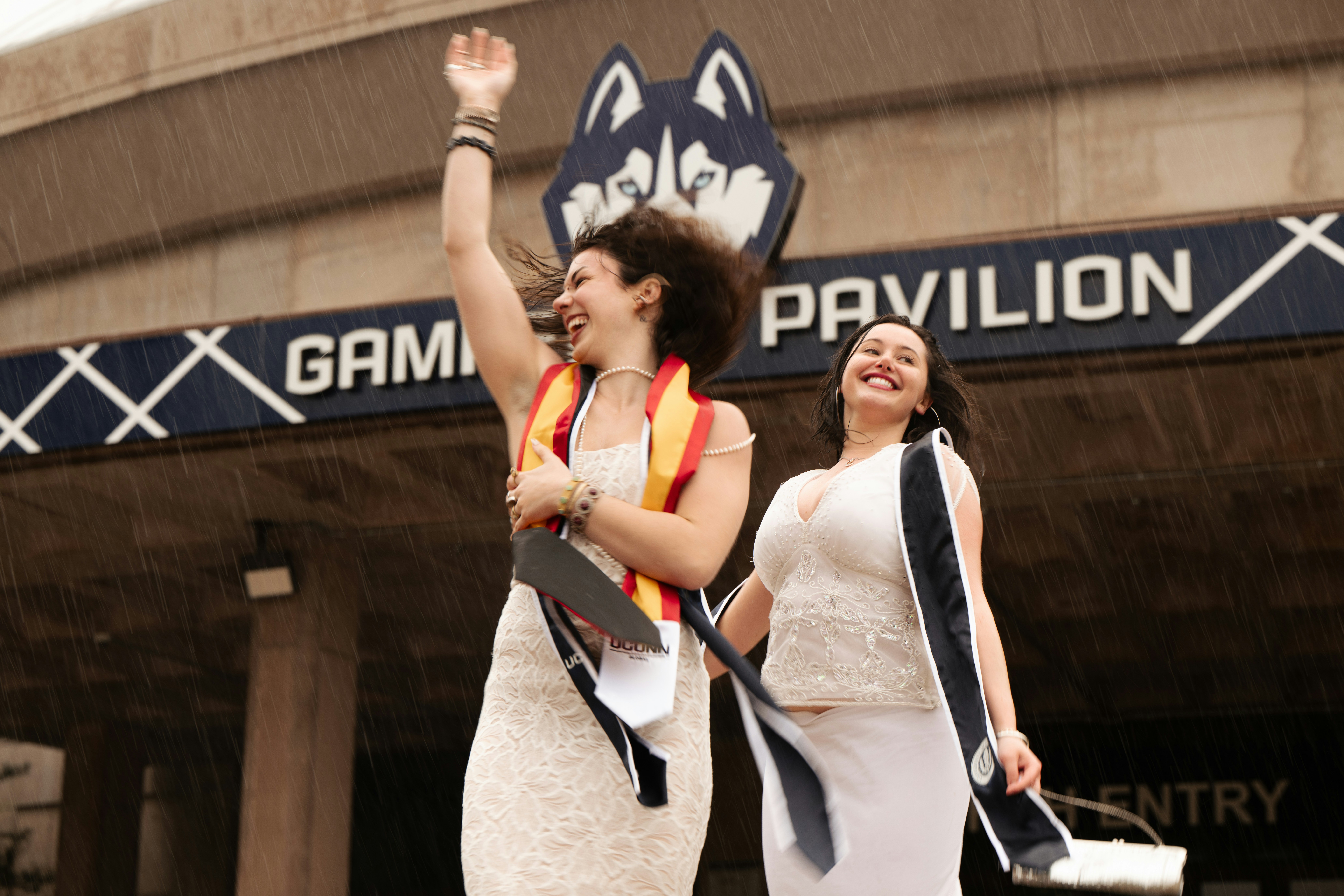 Two women in graduation attire waving happily