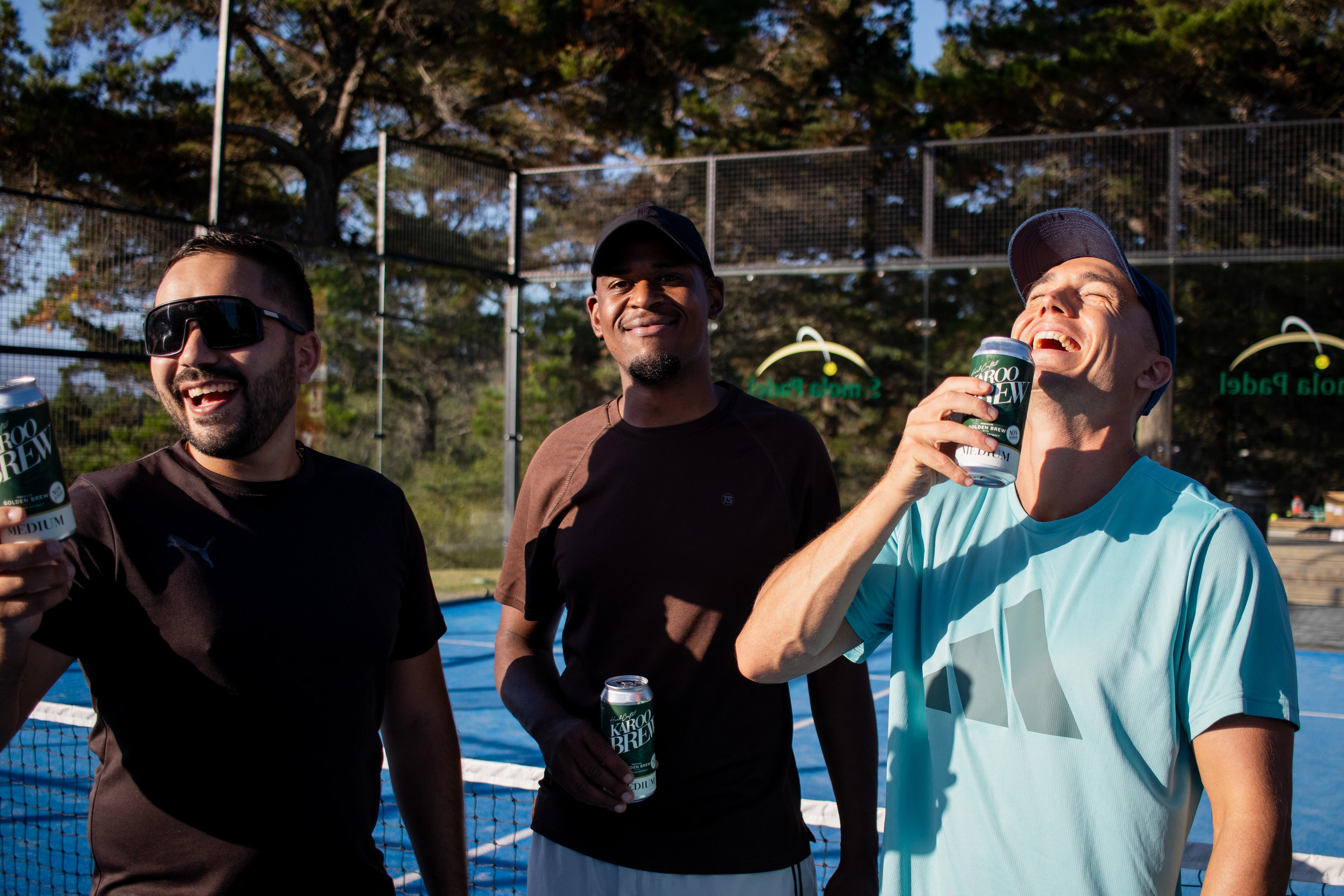 Three men laugh with drinks in hand beside the net of an outdoor padel court in low evening sun