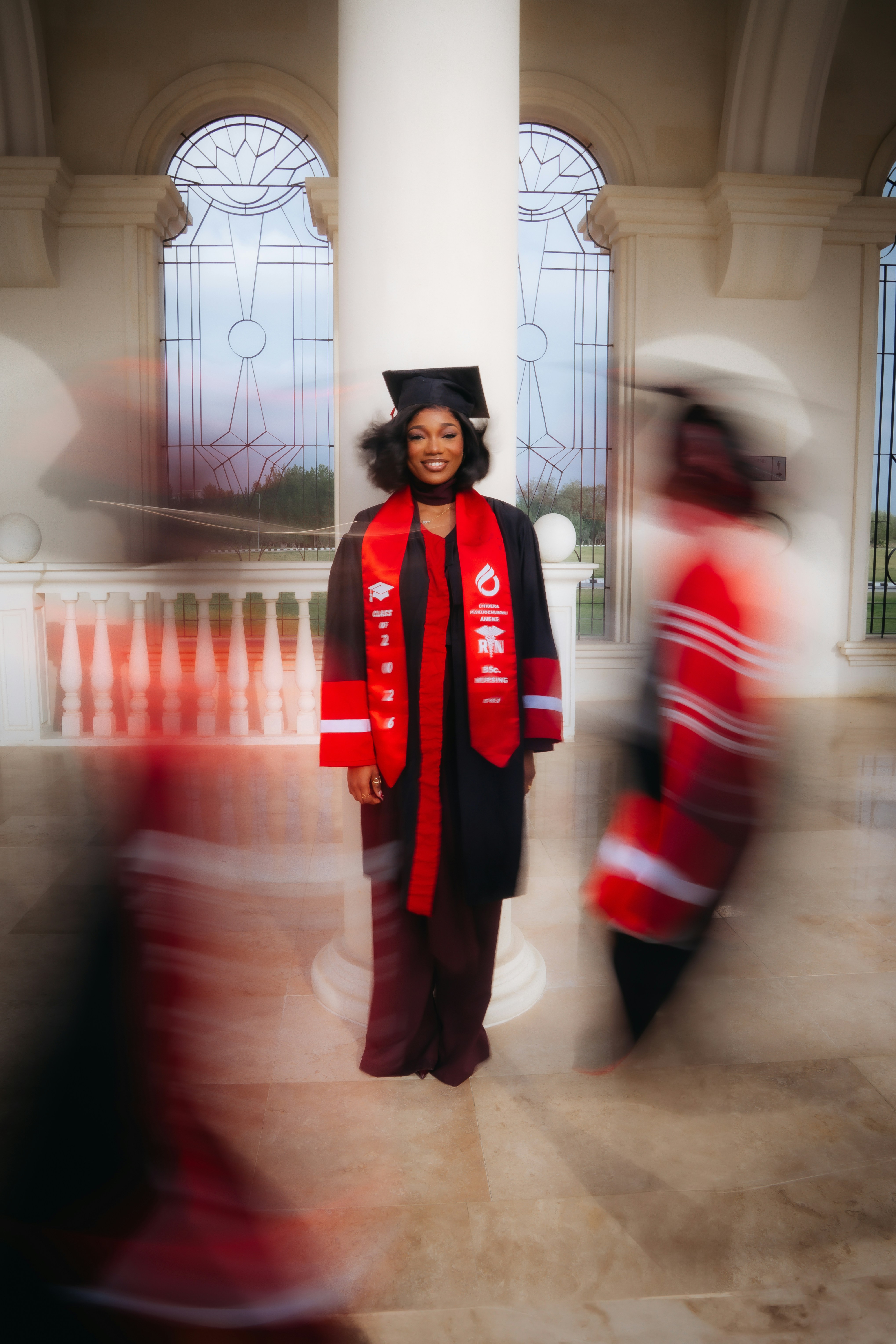 A graduate in regalia stands by a column.