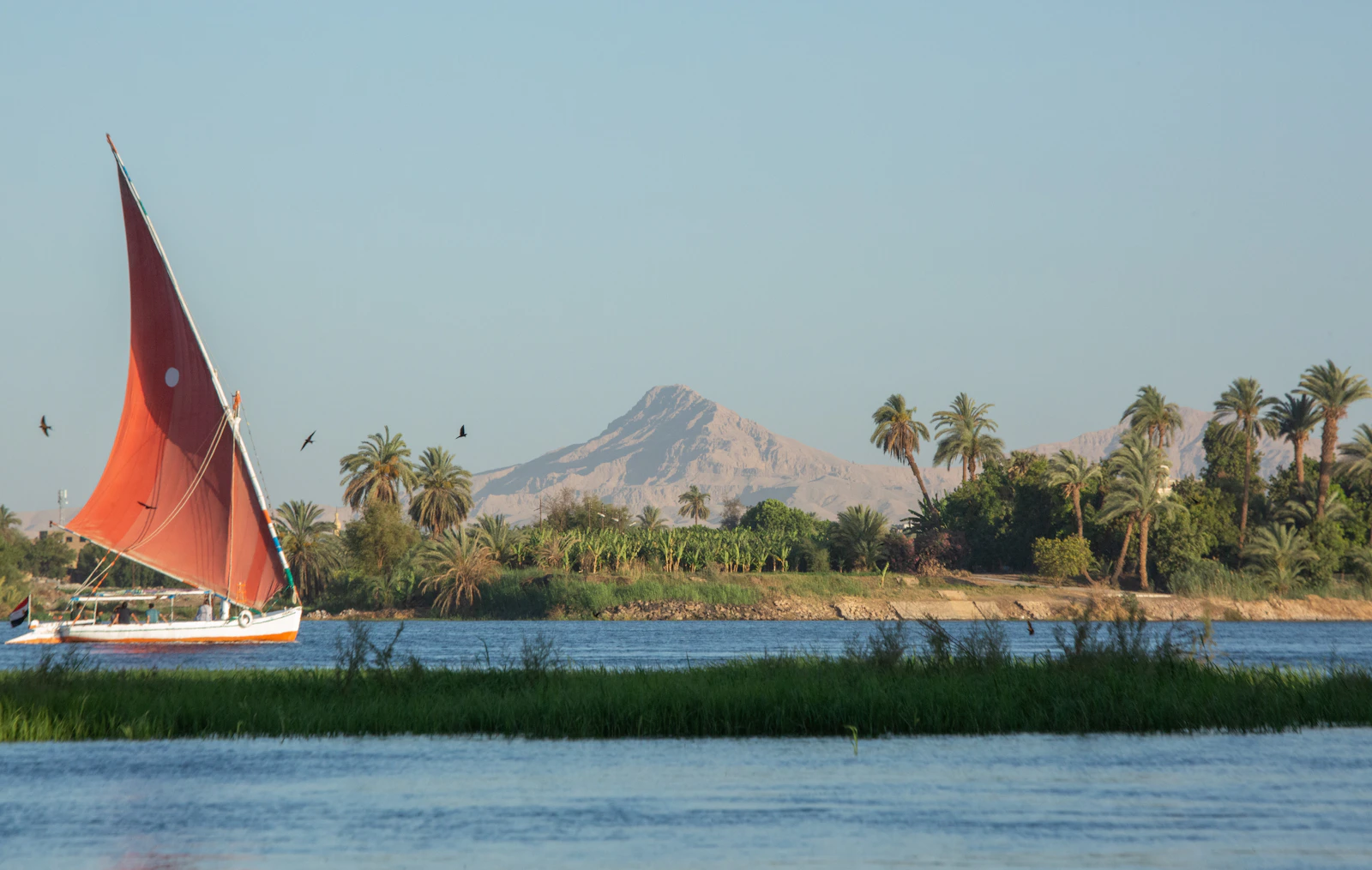 Feluque à voile rouge sur le Nil, entre palmiers et montagnes près de Louxor