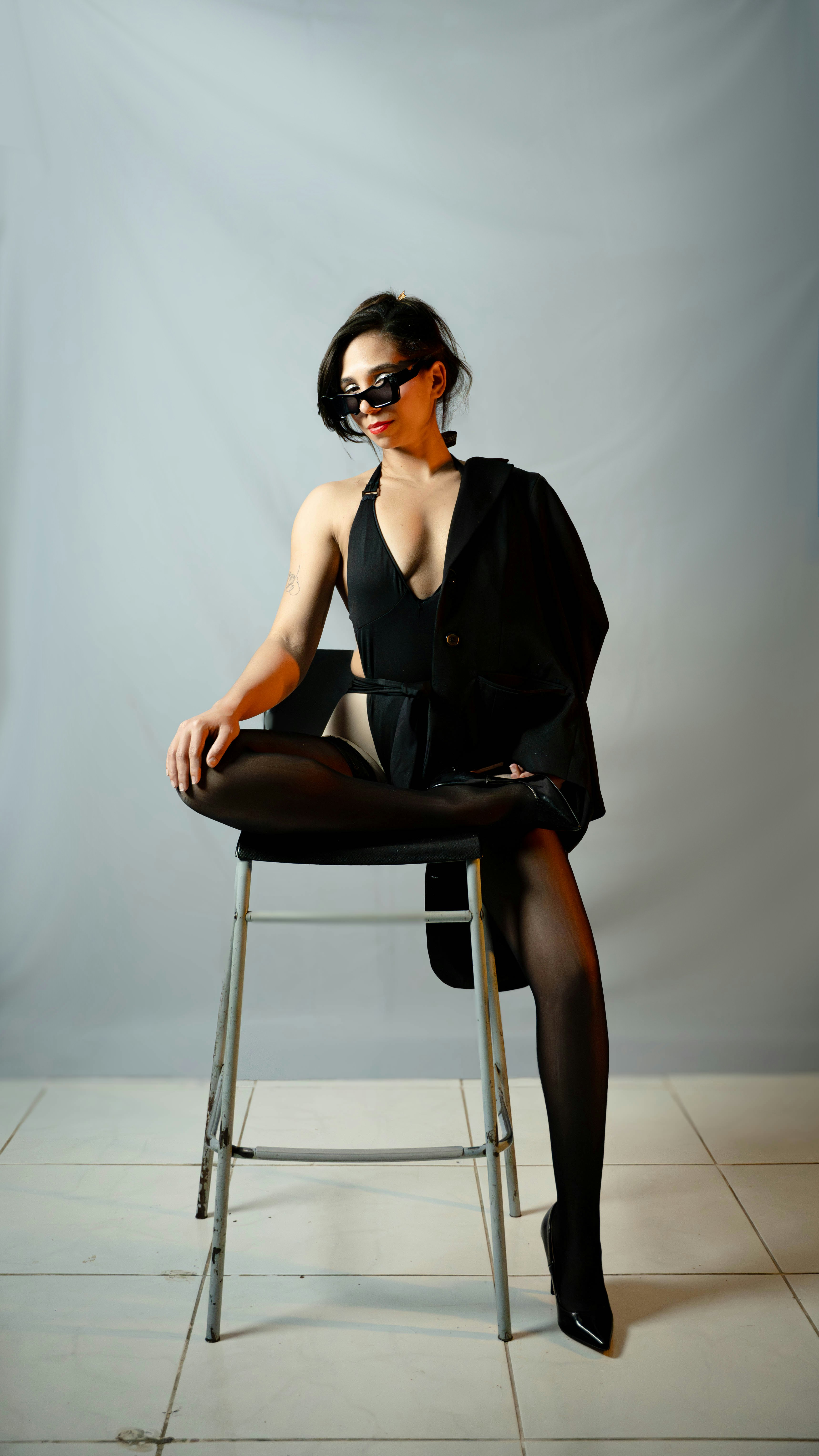 Woman in black swimsuit and blazer sits on stool.
