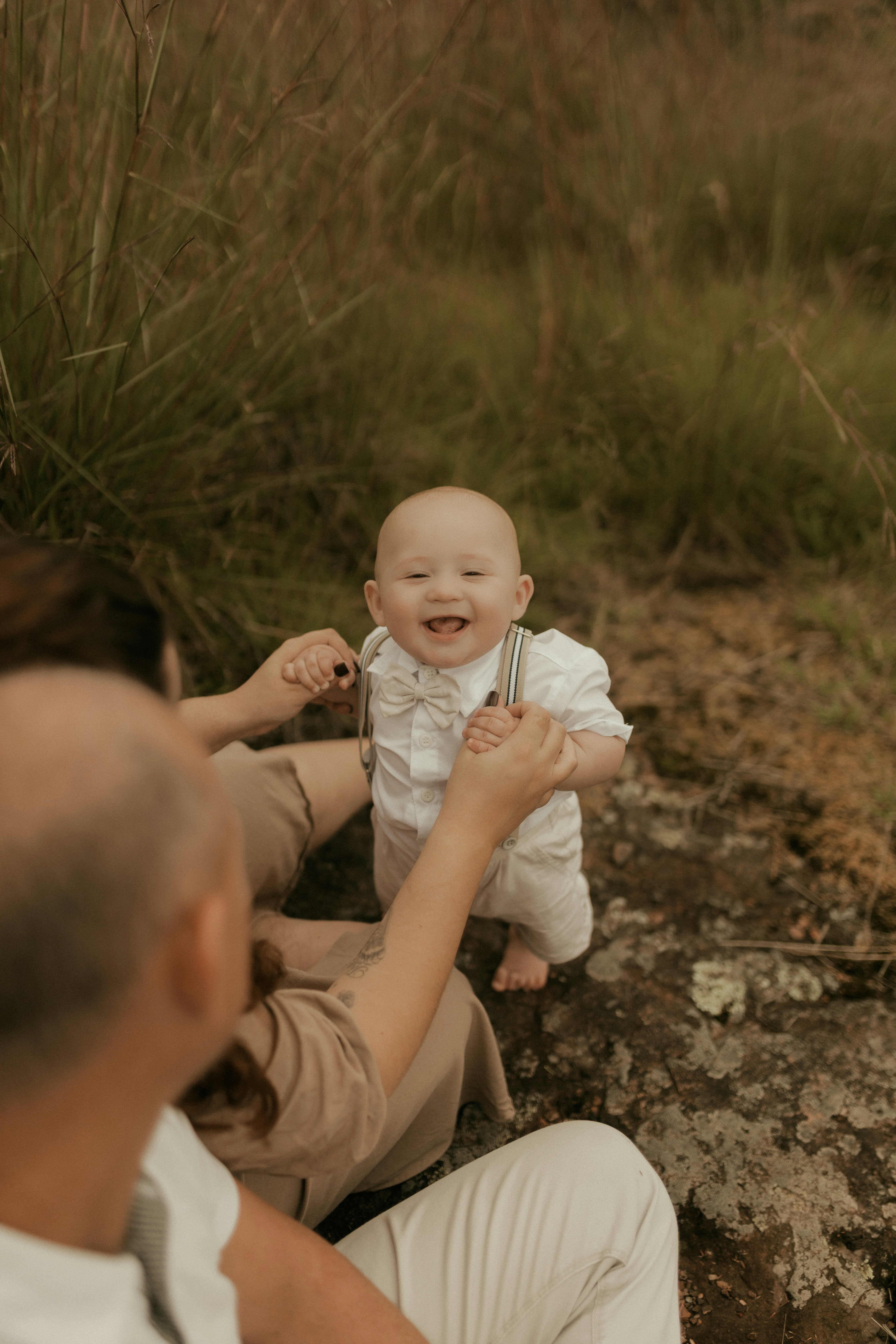 A smiling baby wearing suspenders and a bow tie outdoors.