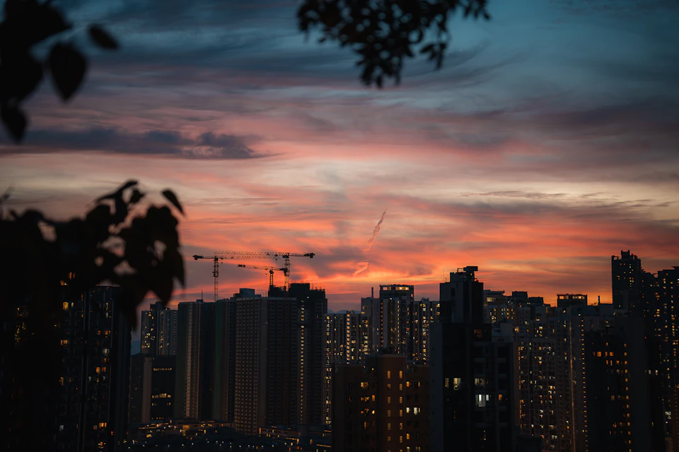 The image depicts a city skyline at sunset, with the sun setting behind the buildings and casting a warm glow over the scene. The sky is painted in shades of orange and pink, creating a stunning backdrop for the urban landscape below. In the foreground, there are two tall buildings standing side by side, one on each side of the frame. The cityscape features numerous other buildings of varying heights, with some appearing closer to the viewer than others due to perspective.

In addition to the buildings, there is a crane visible in the image, suggesting ongoing construction or development in the area. The sky above is filled with clouds, adding depth and texture to the scene.