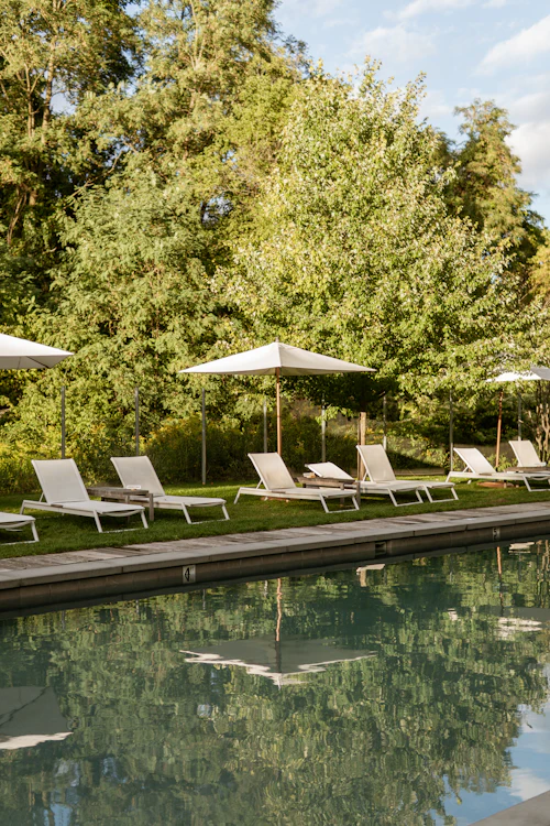 Transats et parasols blancs sur la pelouse, au bord d'une piscine où se reflètent les arbres