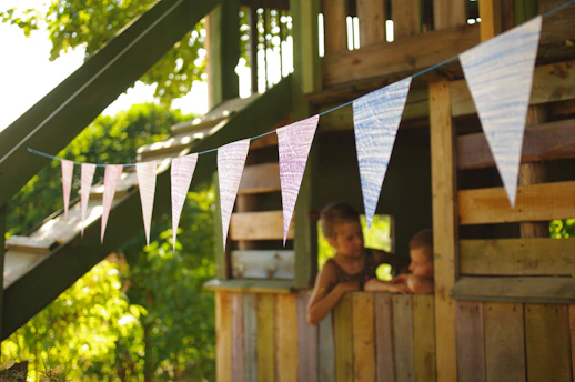 Children peek out from a wooden treehouse decorated with bunting.