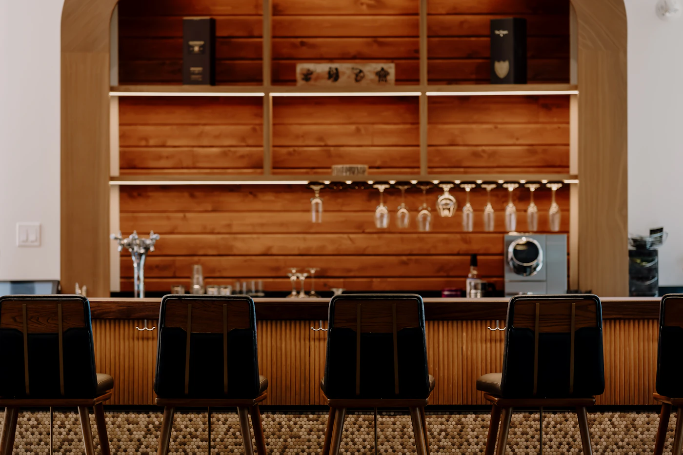 A home bar with wood slat shelving, hanging glasses and a row of dark stools