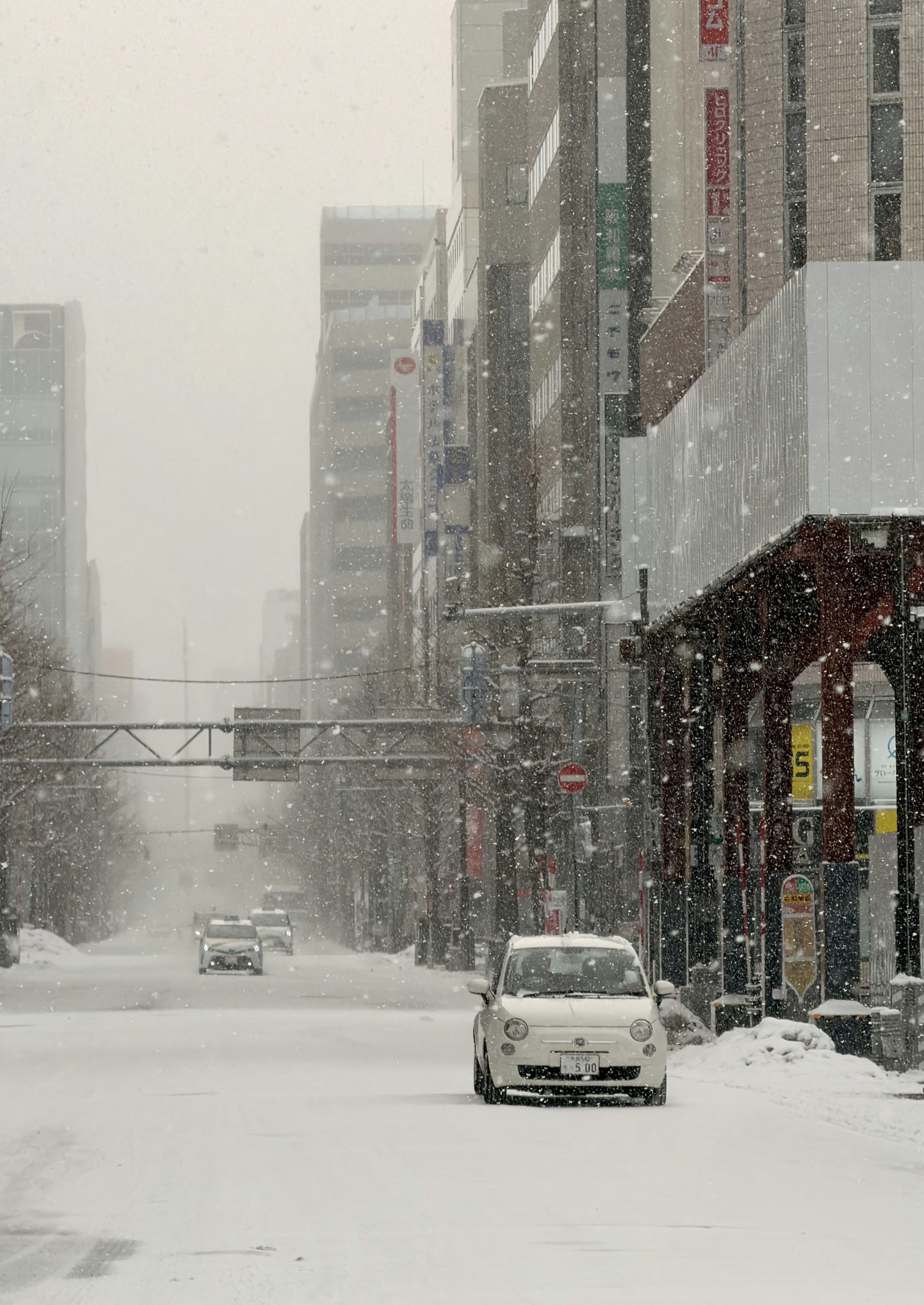 Cars driving on a snowy city street with tall buildings.