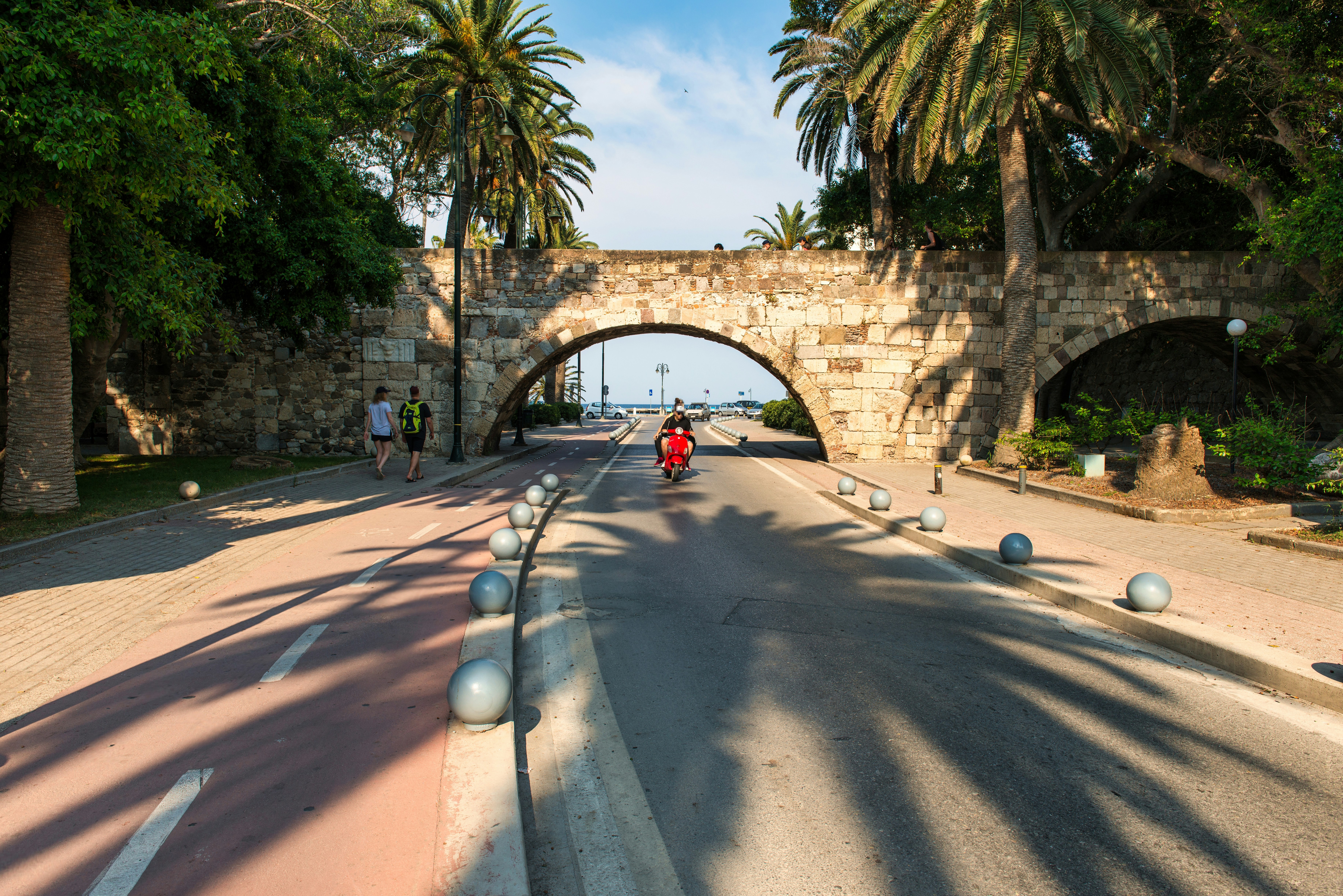 A cyclist rides under a stone bridge with palm trees.