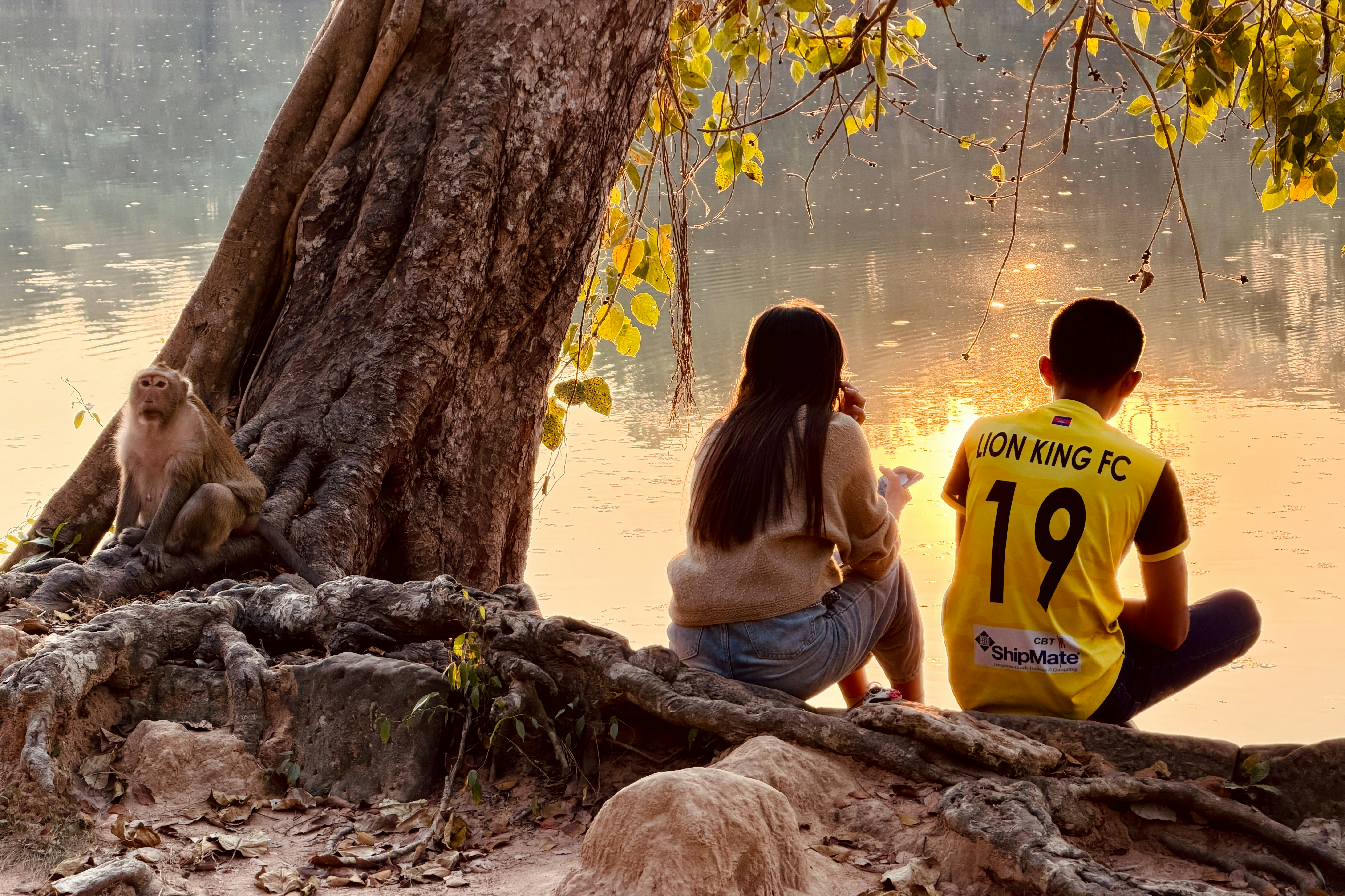 Zwei Menschen und ein Affe an einem See bei Sonnenuntergang.