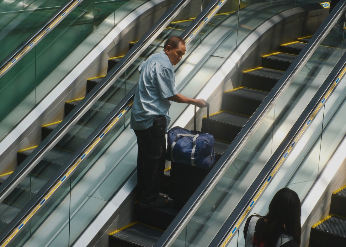A middle-aged man with a hard-sided suitcase on an airport escalator, going up alone, slightly nervous and entirely committed