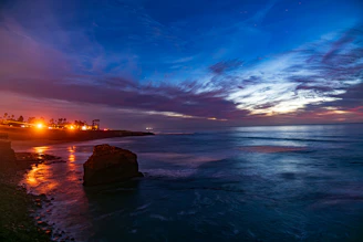 Coastal town lights reflect on ocean at twilight.