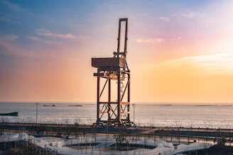 A large industrial crane stands by the ocean at sunset