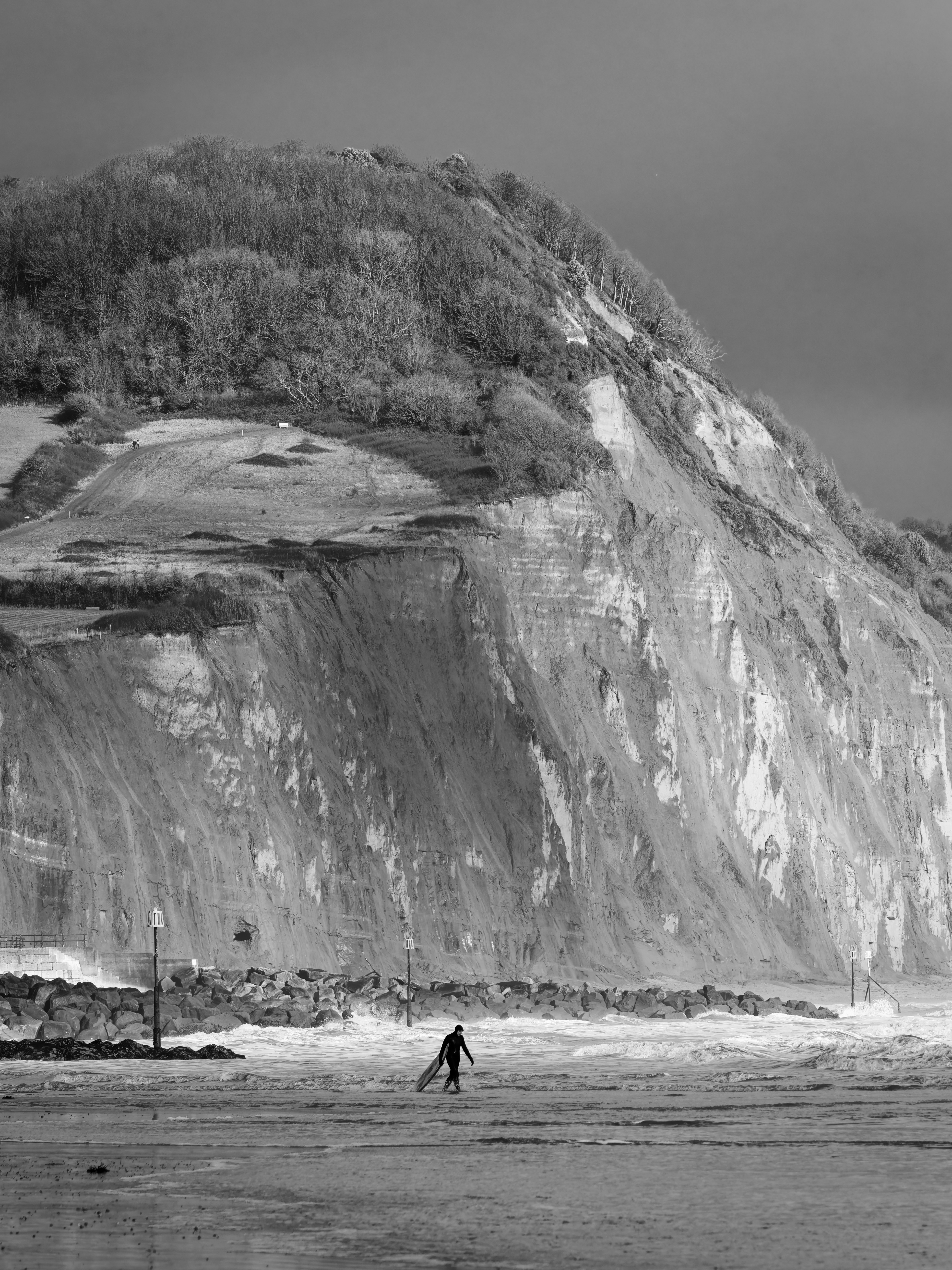 Surfer walks on beach with surfboard near cliffs.