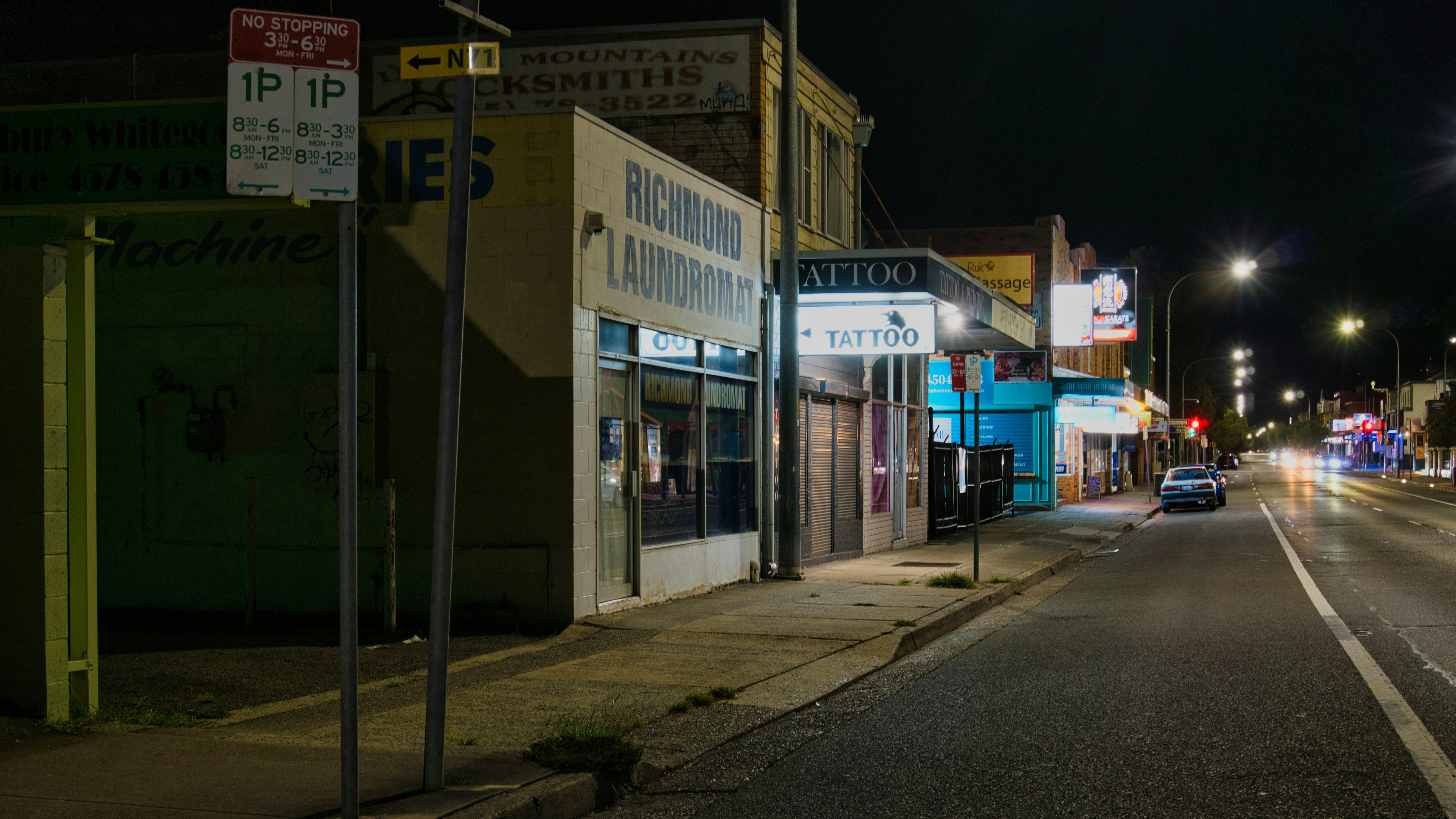 Street scene at night with illuminated storefronts and car lights.