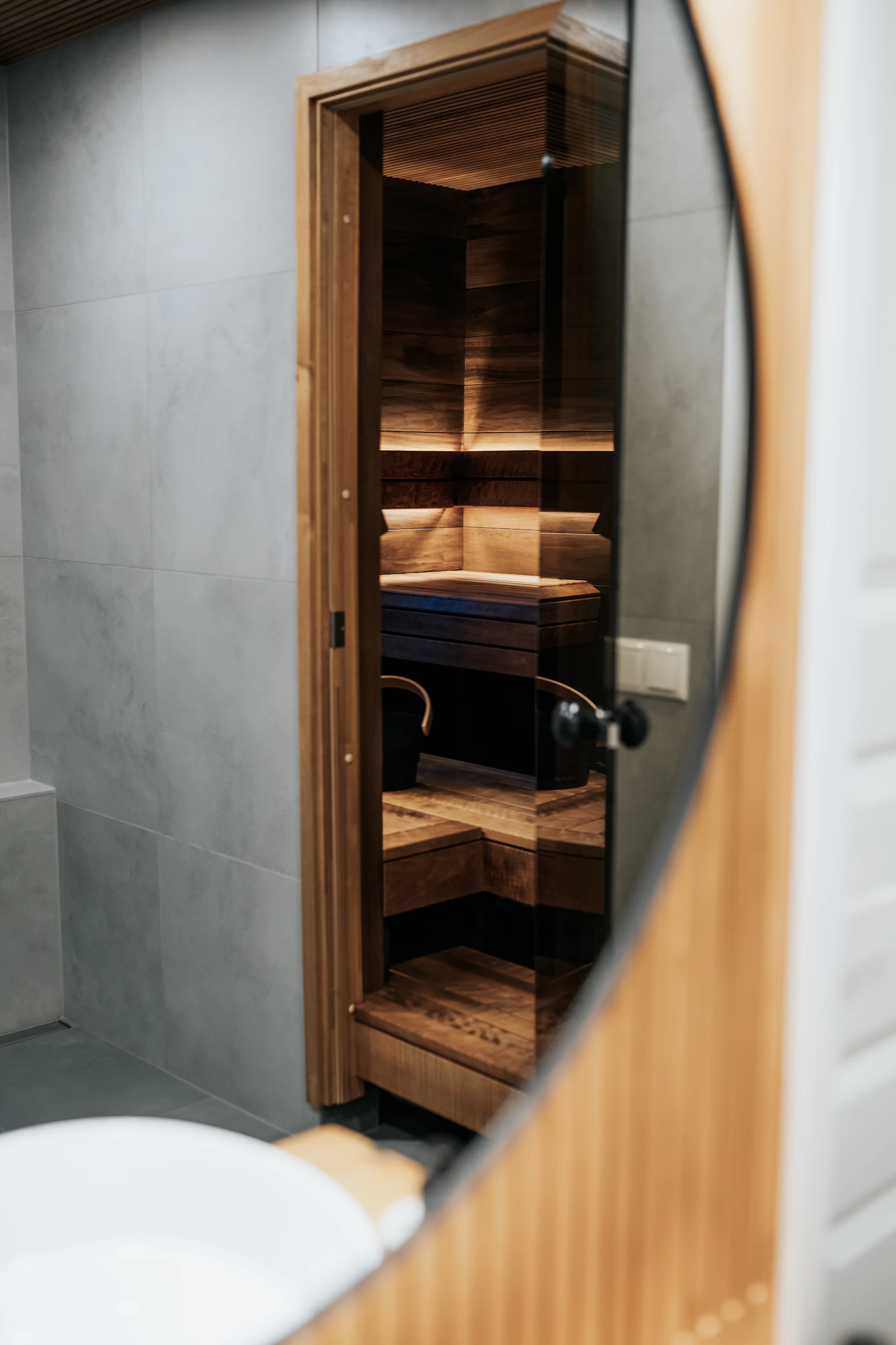 Compact wooden glass-door sauna reflected in a round mirror in a modern bathroom, photographed by Auroom Wellness; illustrative interior, not a specific sauna model.