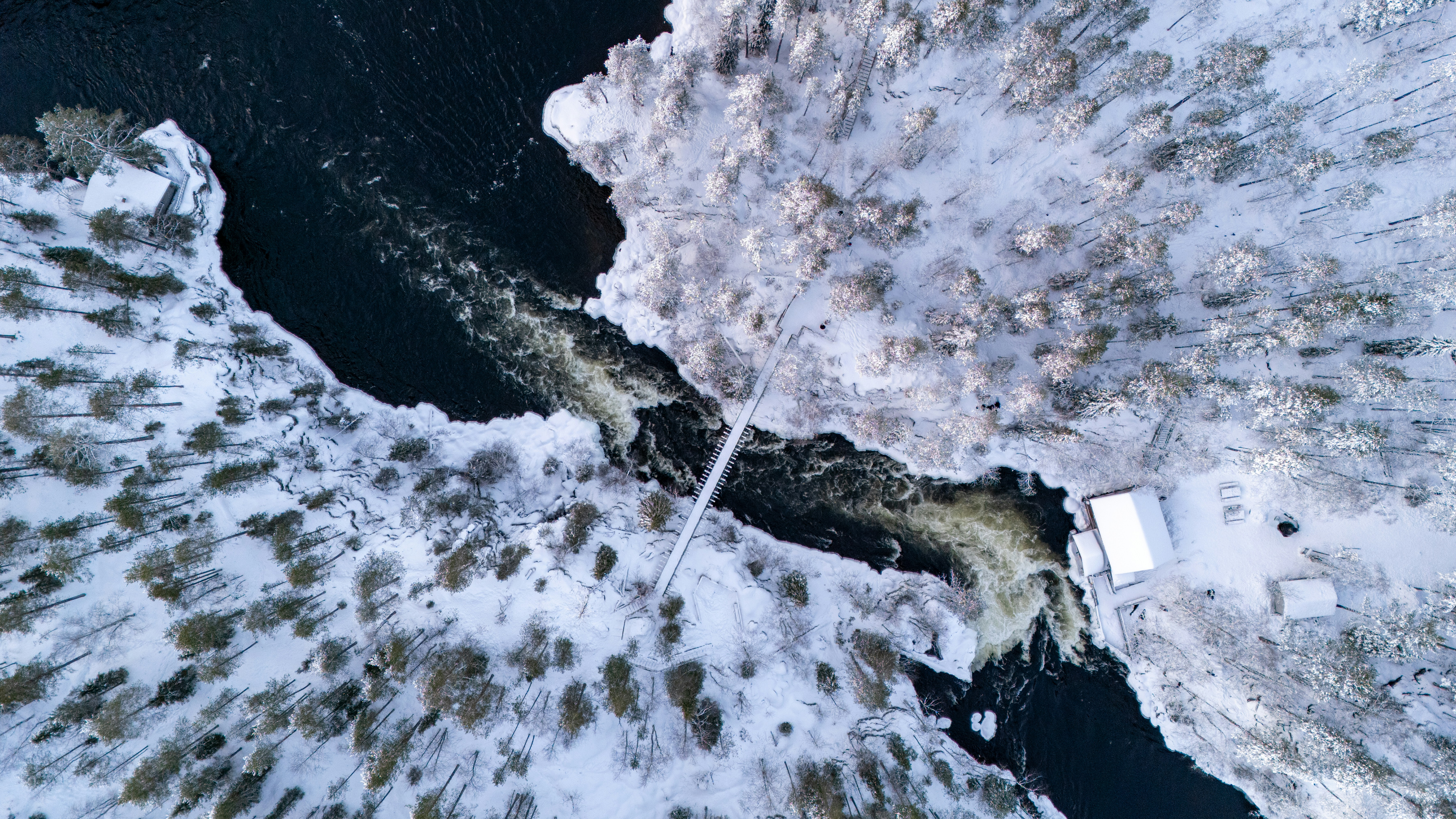 Schneebedeckter Wald umgibt einen rauschenden Fluss mit einem kleinen Gebäude.