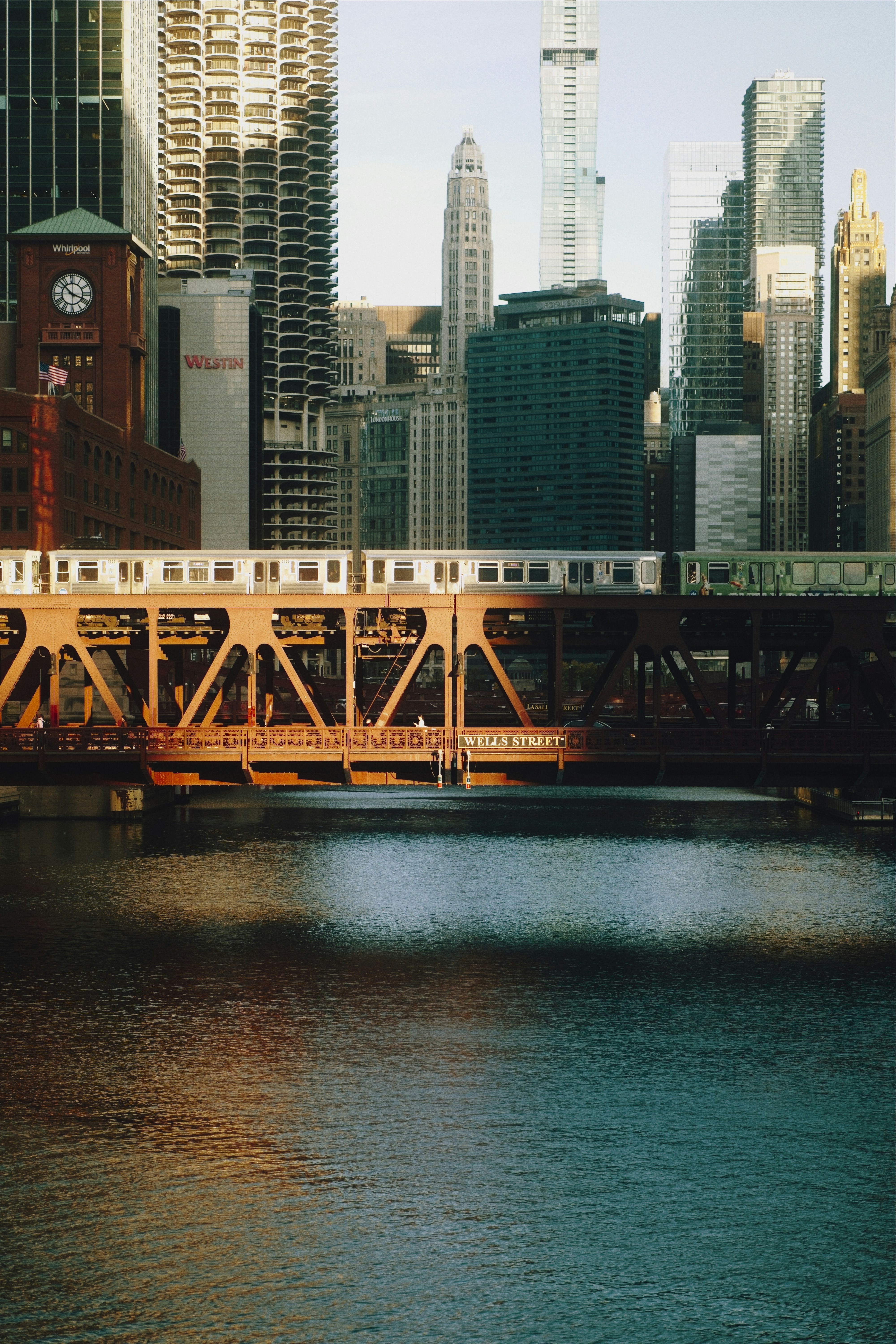 Train crosses a bridge over a river with city buildings.