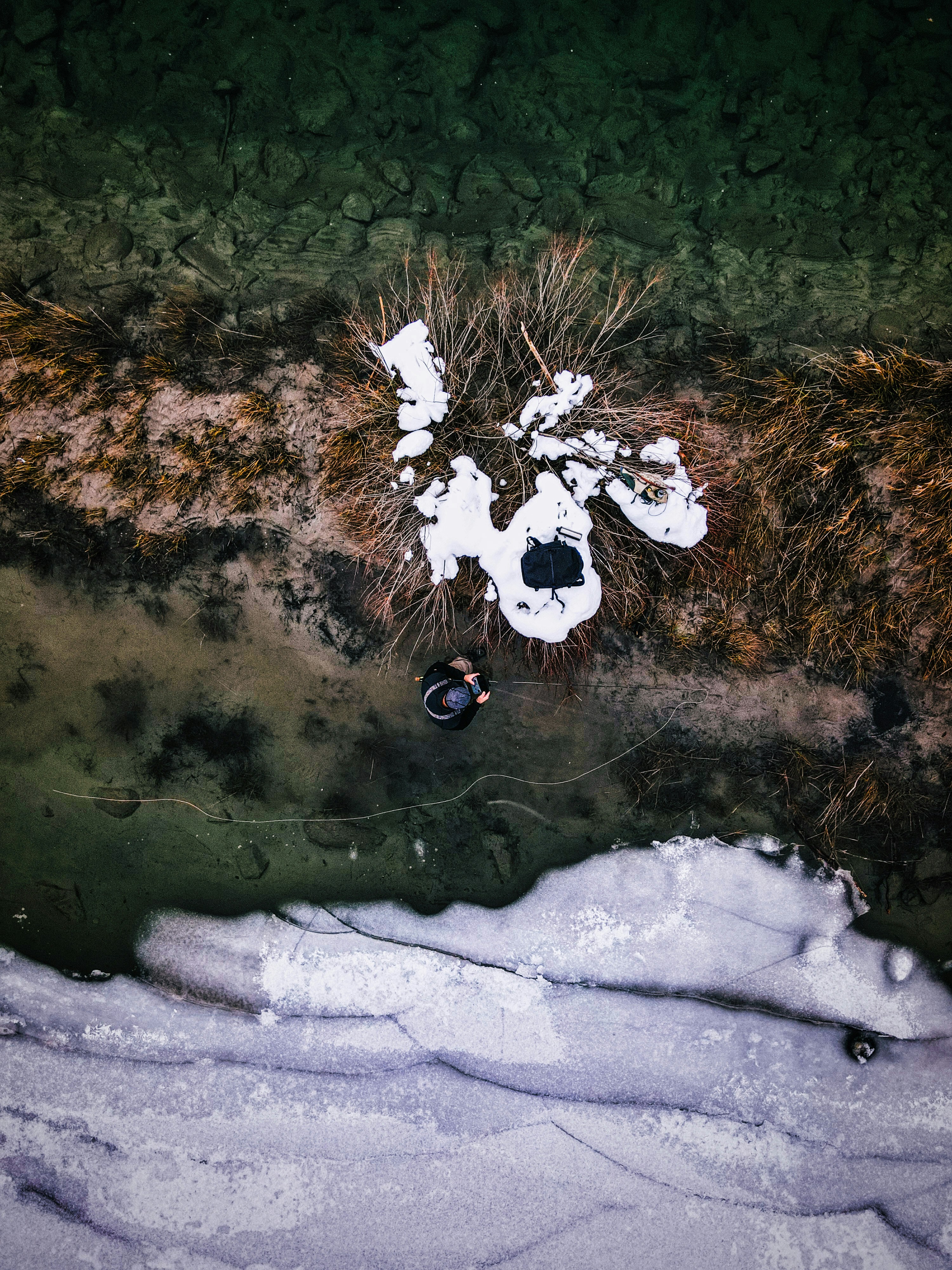 Person fishing by icy water with dry grass and snow